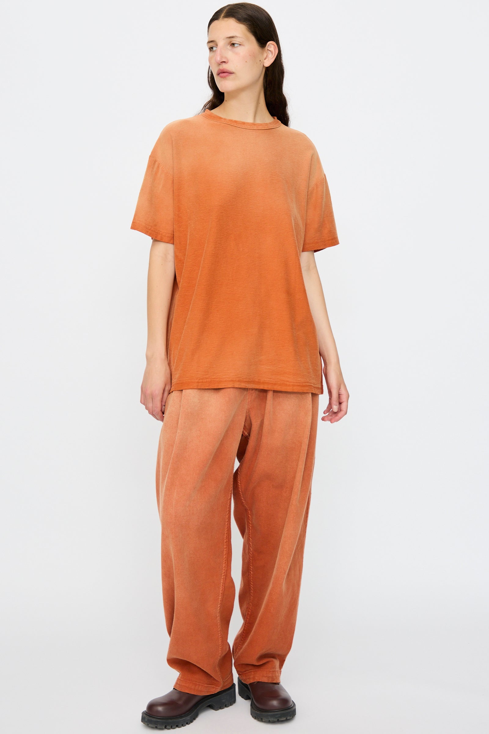 Wearing Dr. Collectors’ Model T. Sunfaded in Rust—loose fit tee and matching wide-leg pants in vibrant orange recycled cotton—a person pairs them with dark brown shoes, standing against a white background for a subtle vintage look.