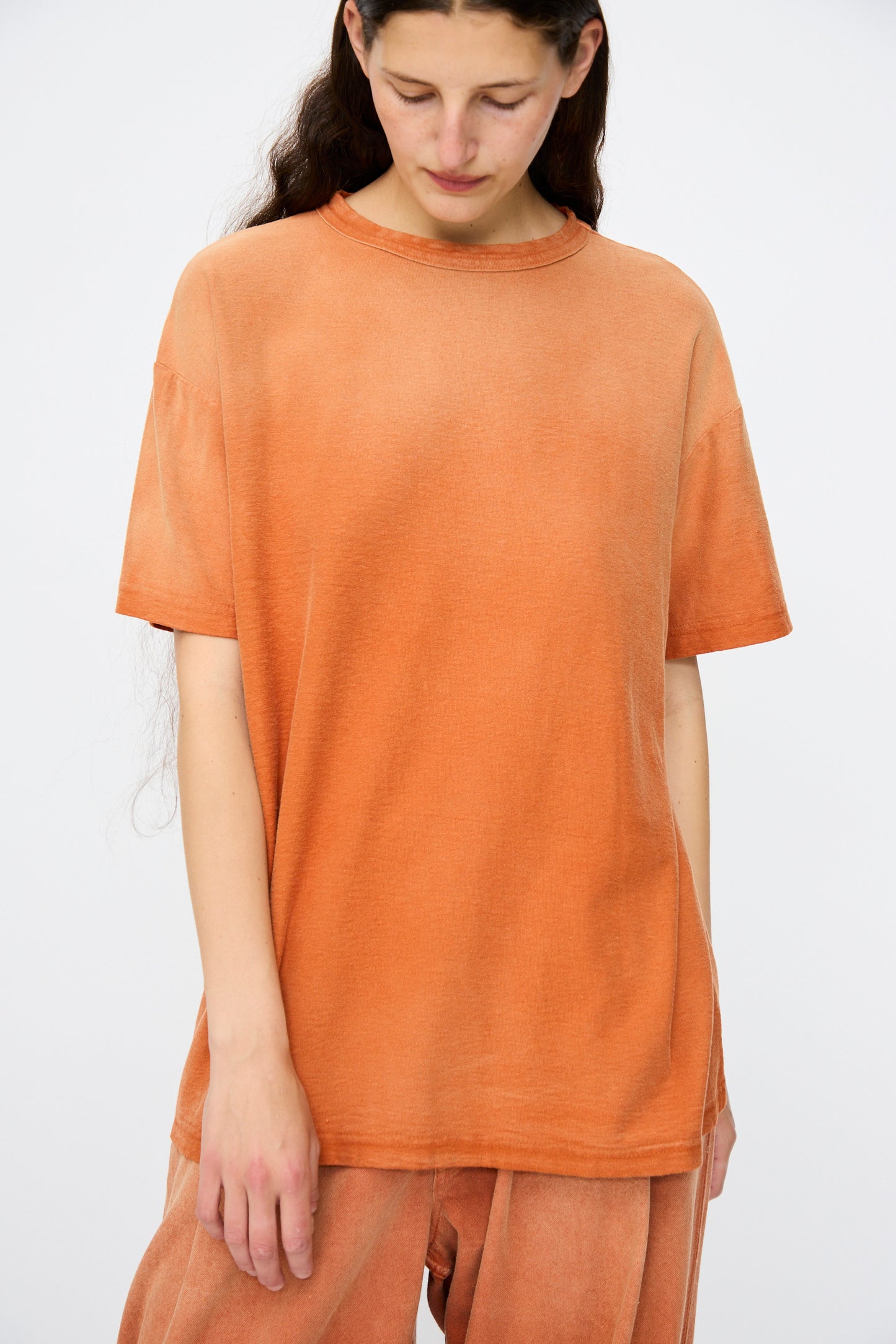 A person with long brown hair wears the Dr. Collectors Model T. Sunfaded in Rust loose fit set, standing against a plain white background and looking downward.