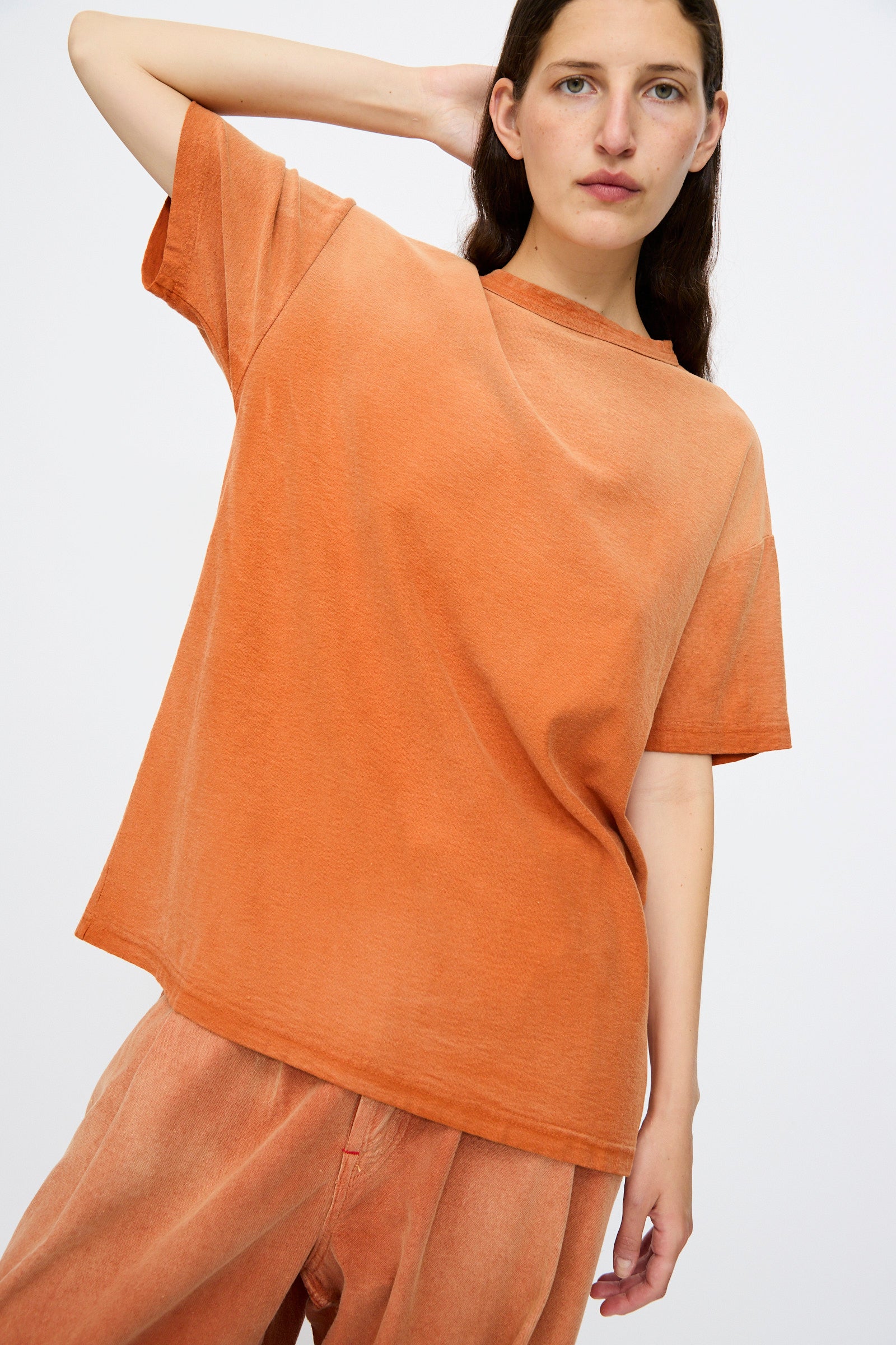 Wearing Dr. Collectors’ Model T. Sunfaded in Rust, a person with long brown hair poses against a plain light background in loose fit recycled cotton tee and matching orange pants.