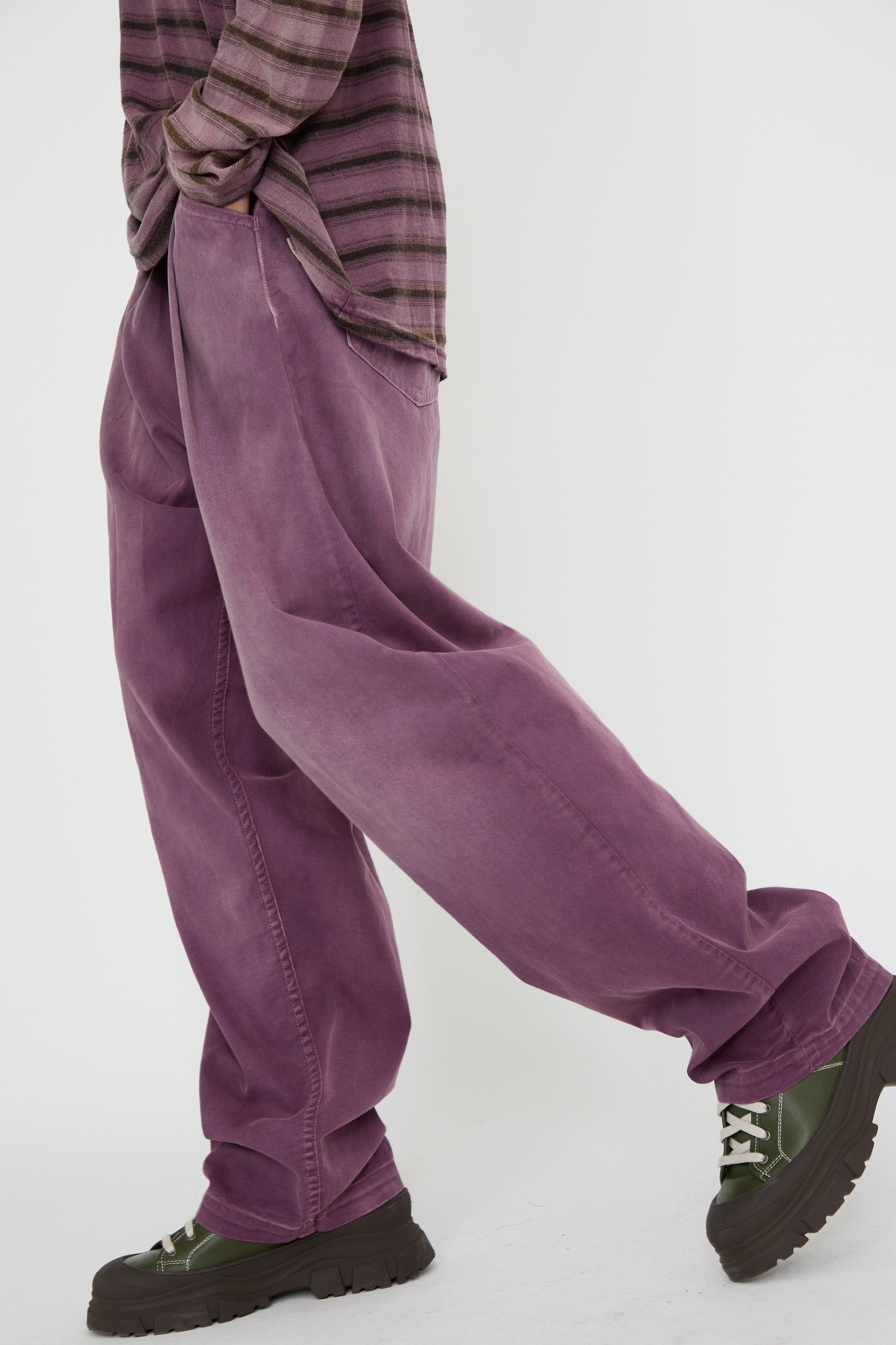 A person wearing a striped shirt, loose straight-leg Dr. Collectors P40 Z Boy's Twill Pant in Sunfaded Mauve, and chunky green shoes walks against a plain backdrop with hands in their pockets. 