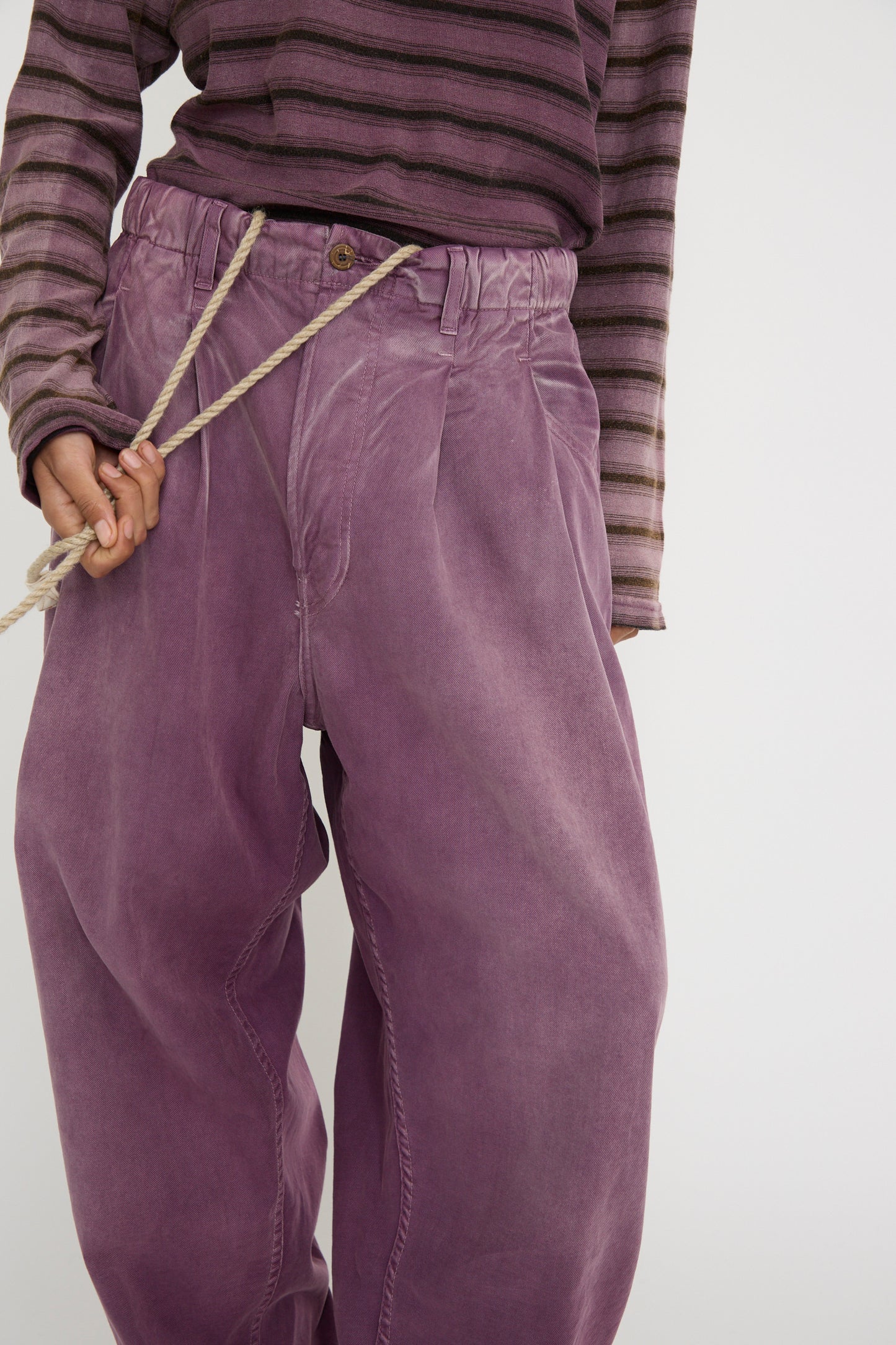 A person wearing a purple striped top and a pair of baggy, oversized P40 Z Boy's Twill Pants in Sunfaded Mauve by Dr. Collectors, holding the twill drawstrings against a white background. 