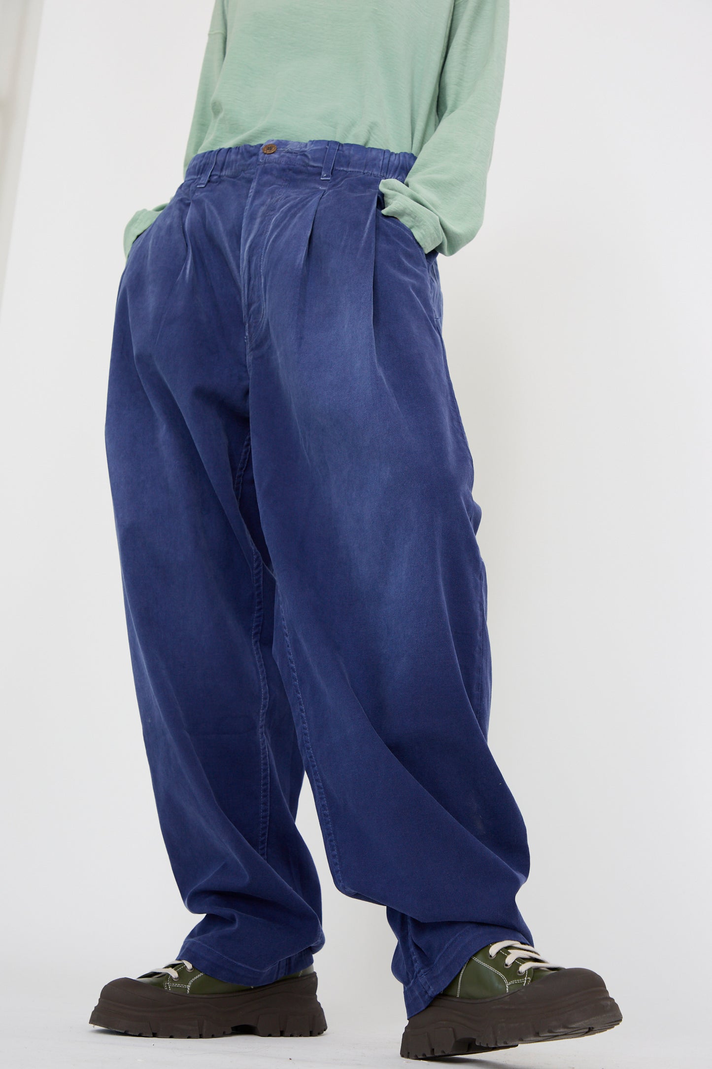 A person wearing a light green top, oversized P40 Z Boy's Twill Pant in Sunfaded Sailor Blue from Dr. Collectors, and dark chunky shoes. Hands tucked into pant pockets against a plain white background.