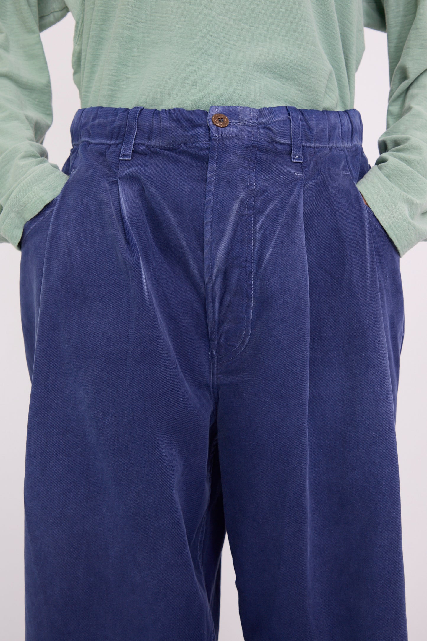 A close-up shot of a person dressed in a green shirt and P40 Z Boy's Twill Pant in Sunfaded Sailor Blue by Dr. Collectors, crafted from Japanese cotton, stands casually with hands in pockets. 