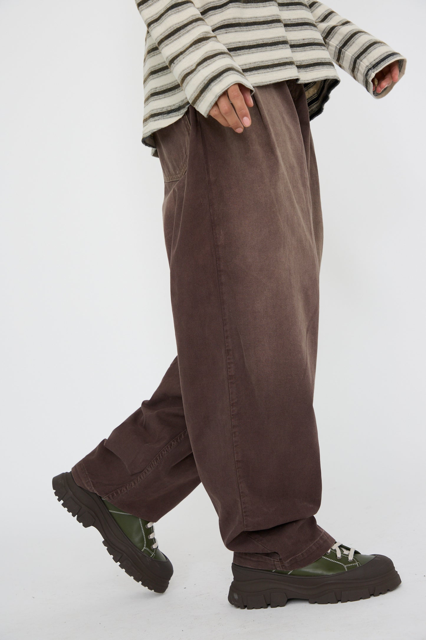 A person is walking, dressed in a striped long-sleeve shirt and oversized P40 Z Boy's Twill Pant Sunfaded in Cappuccino by Dr. Collectors, crafted from soft Japanese cotton with an elasticated waistband. Complementing the look are green shoes set against a plain background.