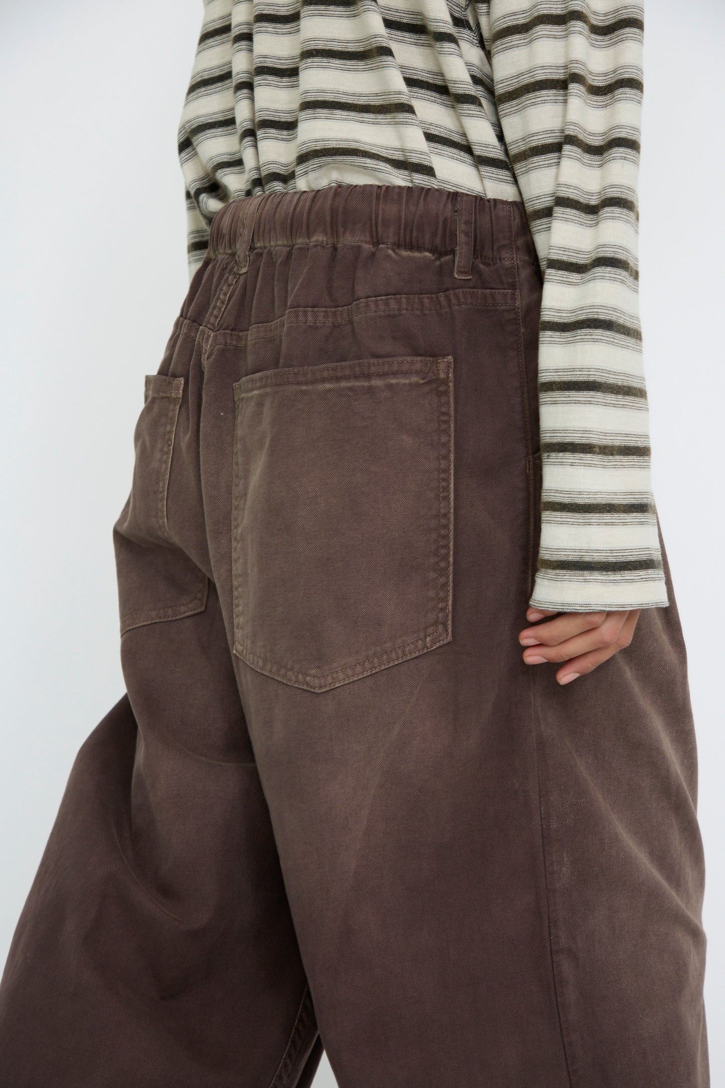 A close-up of a person from the b ack standing against a plain background wearing Dr. Collectors' P40 Z Boy's Twill Pant Sunfaded in Cappuccino, featuring an elasticated waistband and visible back pockets, paired with a striped shirt.