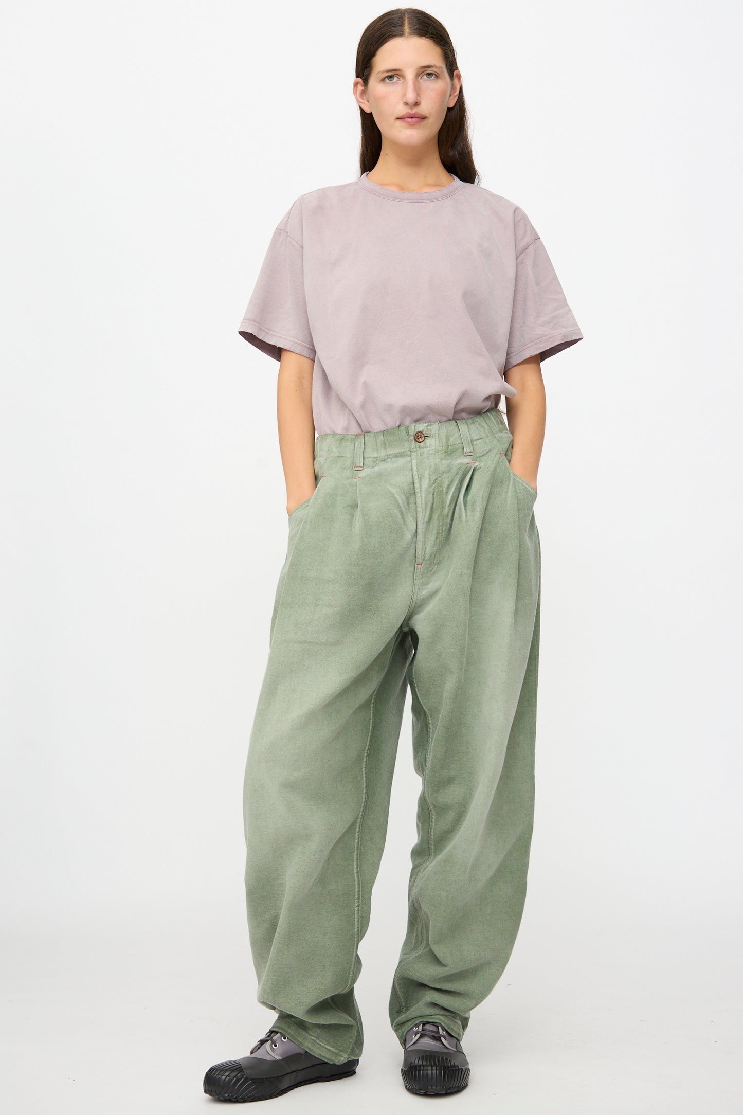 A person stands against a white background, wearing a loose lavender T-shirt made from soft Japanese cotton, Dr. Collectors P40 Z Boys Pant in Sunfaded Army Green featuring a relaxed fit silhouette, and black sneakers, with hands in pockets and a neutral expression.