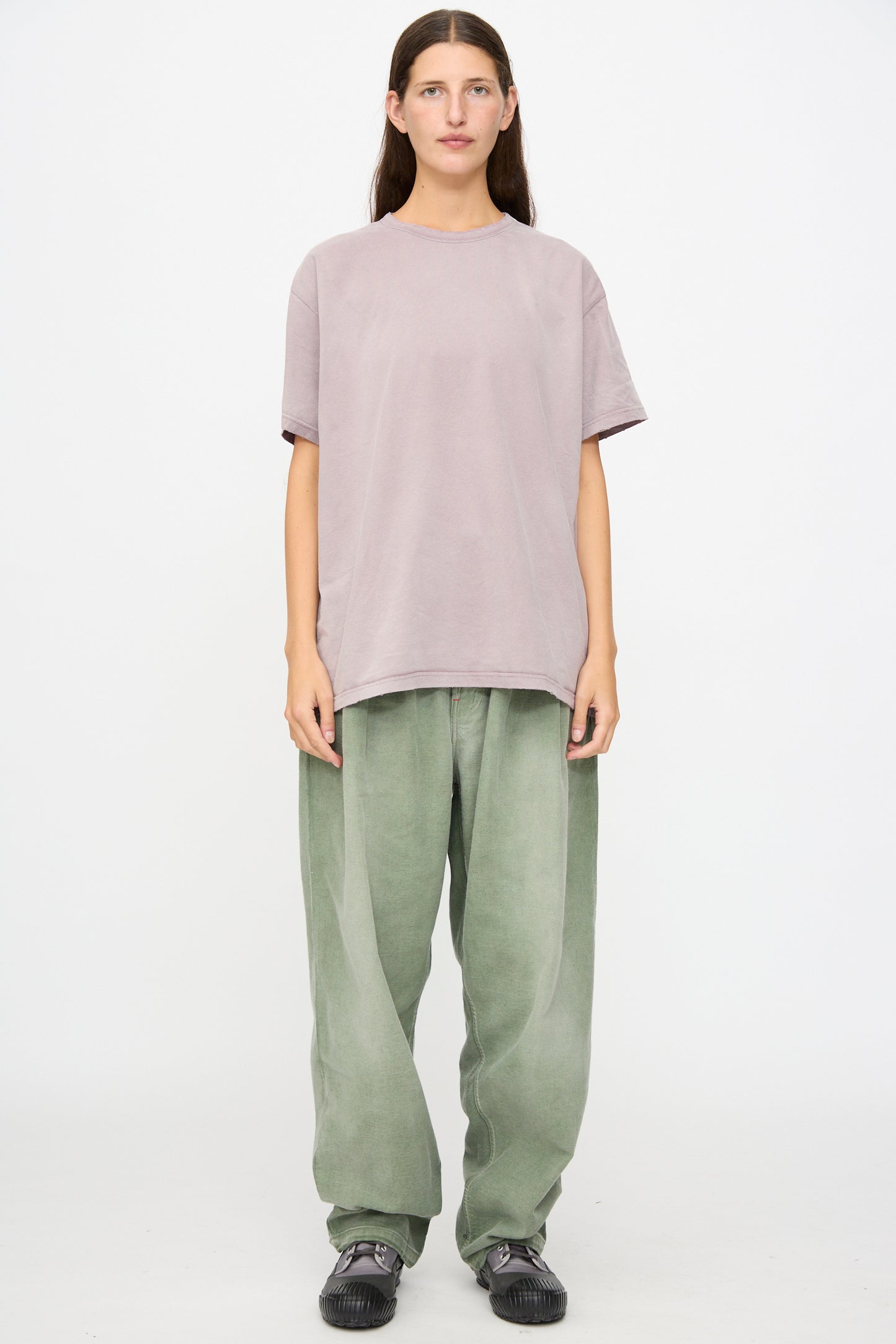 Person standing against a plain white background, wearing a loose light purple t-shirt crafted from soft Japanese cotton, Dr. Collectors P40 Z Boys Pant in Sunfaded Army Green, and black sneakers.