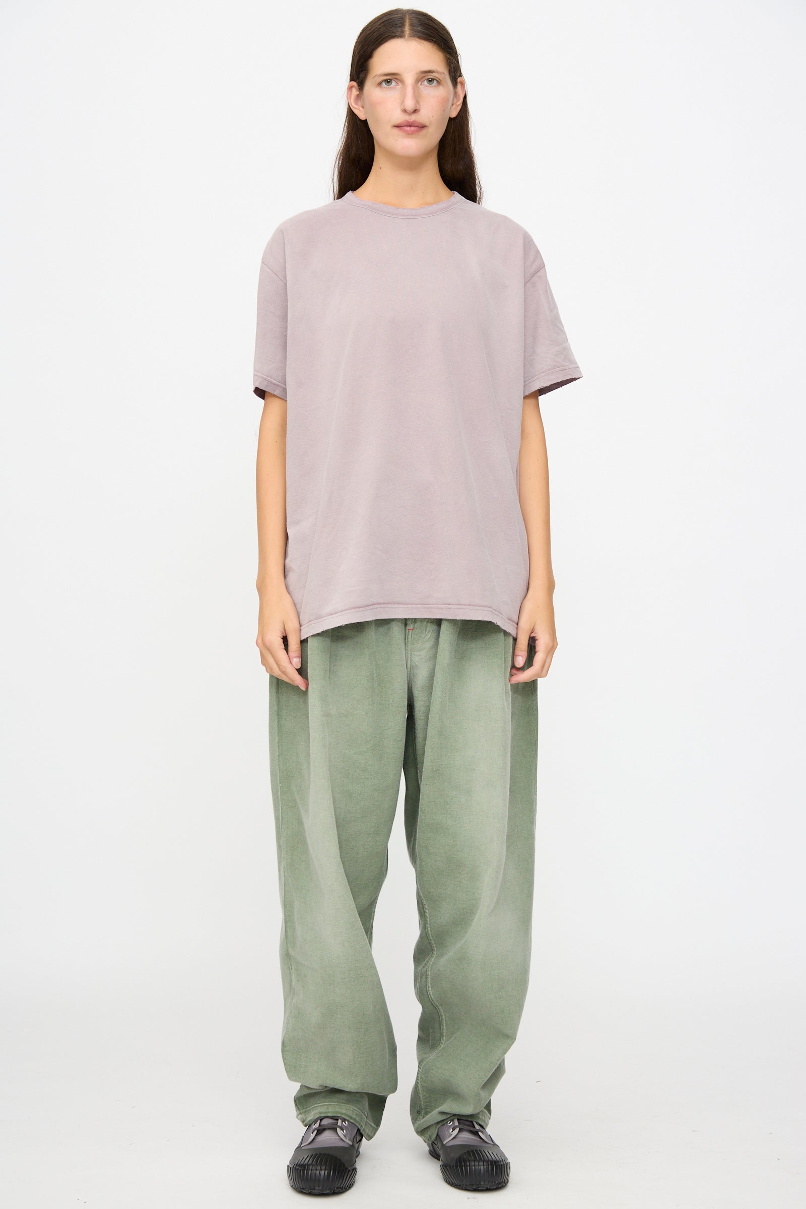 Person standing against a plain white background, wearing a loose light purple t-shirt crafted from soft Japanese cotton, Dr. Collectors P40 Z Boys Pant in Sunfaded Army Green, and black sneakers.
