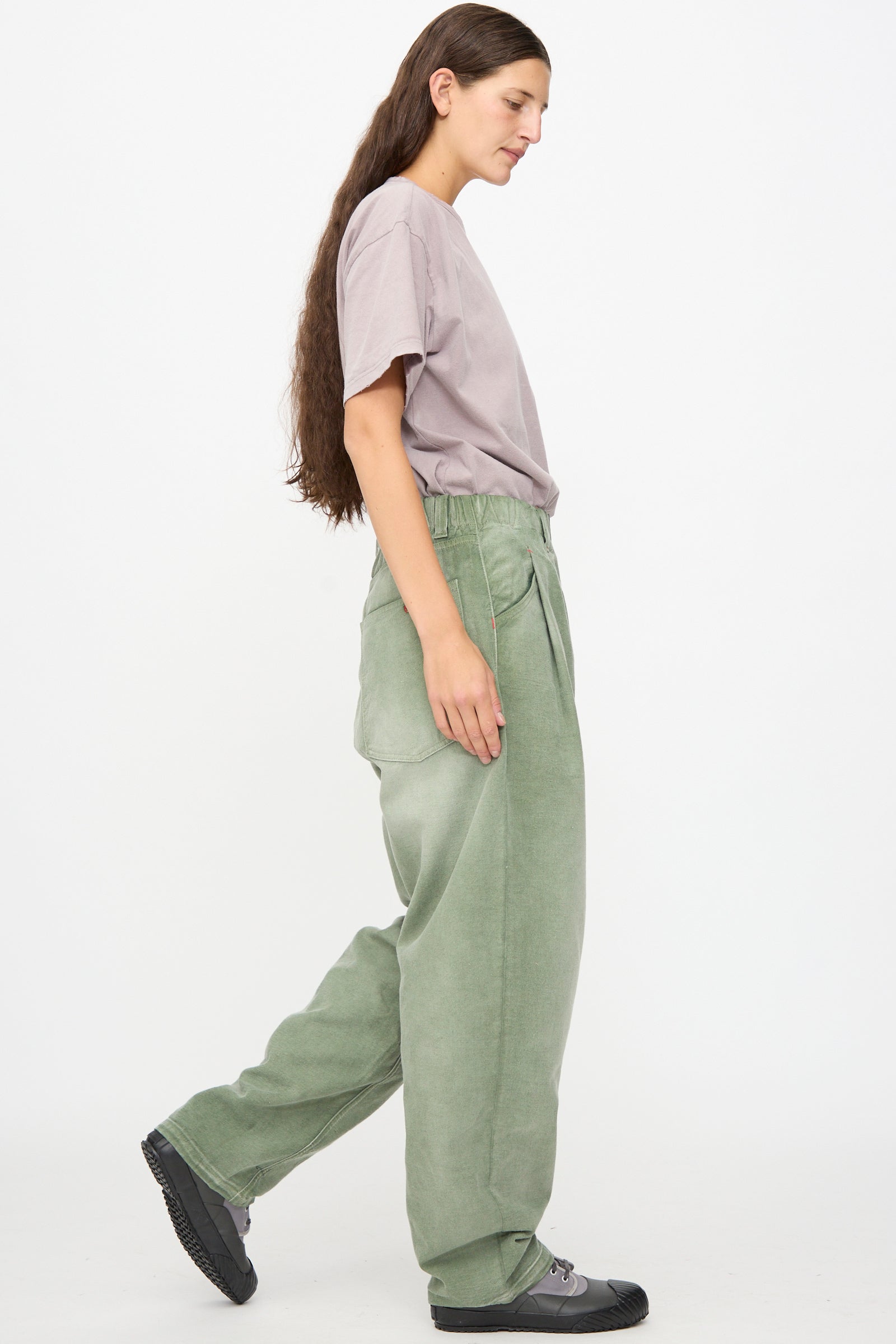 Person with long brown hair wearing a loose lavender t-shirt made from soft Japanese cotton, paired with Dr. Collectors P40 Z Boys Pant in Sunfaded Army Green and black sneakers, standing in profile against a plain white background.