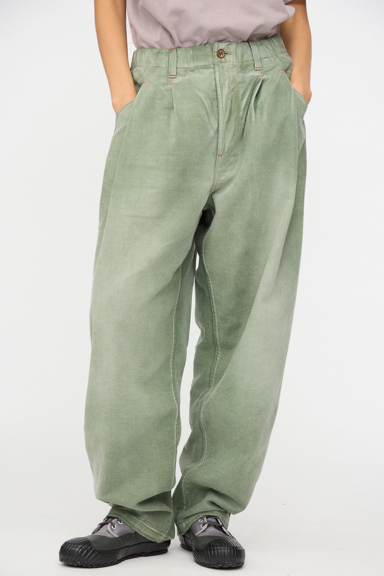 A person wearing Dr. Collectors’ P40 Z Boys Pant in Sunfaded Army Green, featuring a relaxed fit and crafted from soft Japanese cotton, stands against a plain white background with hands in pockets, paired with black shoes and a grey shirt.