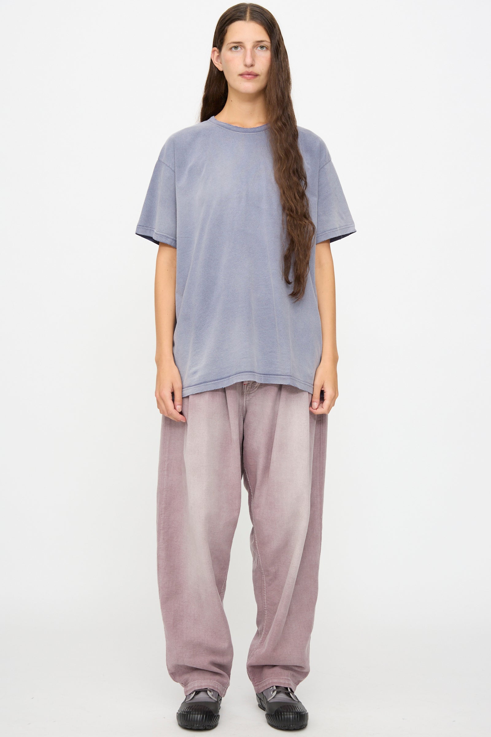 A person with long brown hair down stands against a white background wearing a loose lavender t-shirt, Dr. Collectors P40 Z Boys Pant in Sunfaded Dusty Orchid—a relaxed fit, straight leg pant crafted from light purple Japanese cotton—and black shoes.