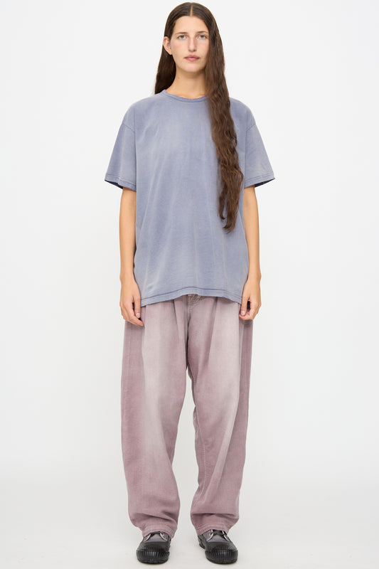 A person with long brown hair down stands against a white background wearing a loose lavender t-shirt, Dr. Collectors P40 Z Boys Pant in Sunfaded Dusty Orchid—a relaxed fit, straight leg pant crafted from light purple Japanese cotton—and black shoes.