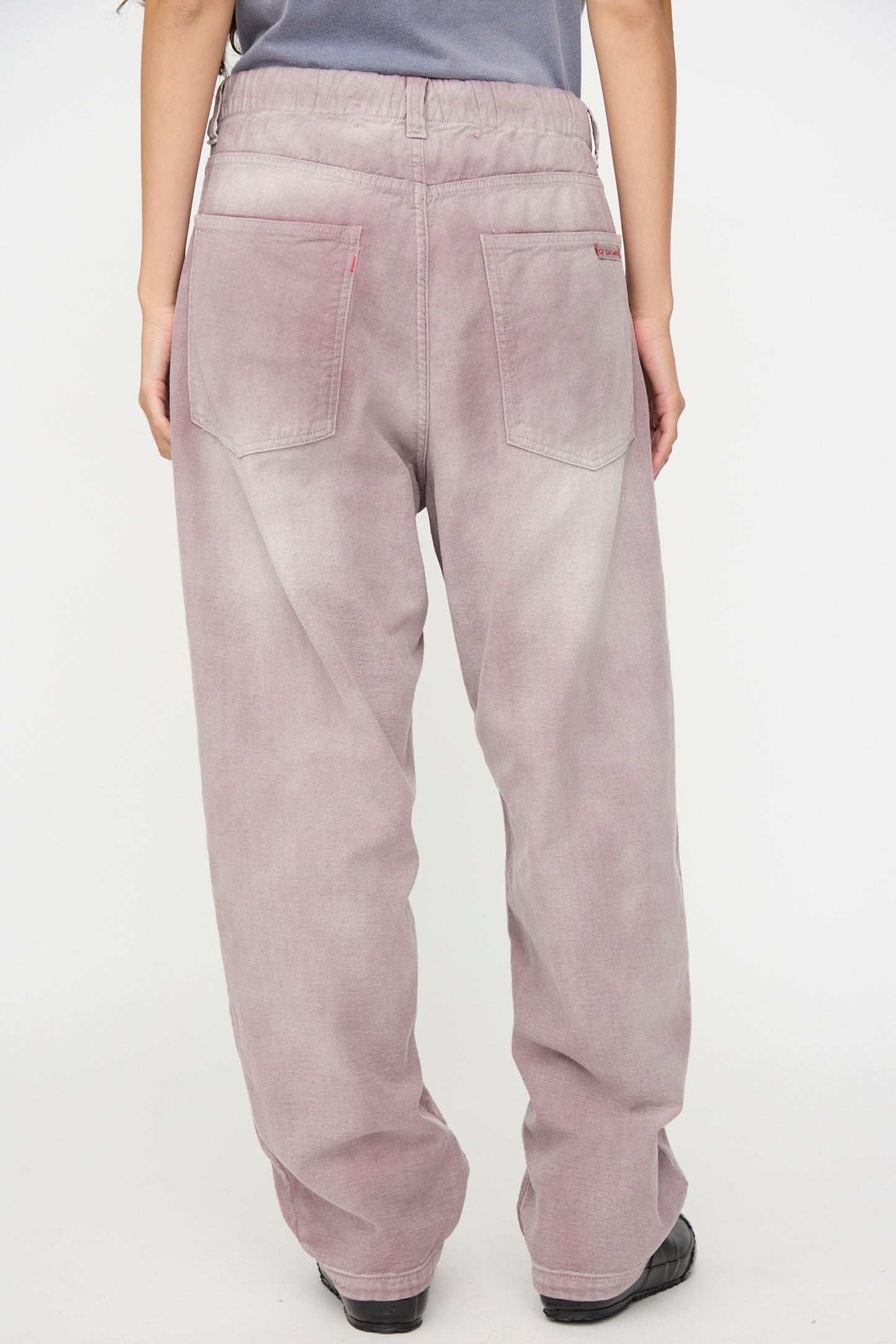 A person is seen from the back wearing Dr. Collectors’ P40 Z Boys Pant in Sunfaded Dusty Orchid—a relaxed fit style featuring a loose straight leg cut, large back pockets, and an elastic waistband, paired with black shoes. The comfortable, faded dusty orchid jeans are perfect for everyday wear.