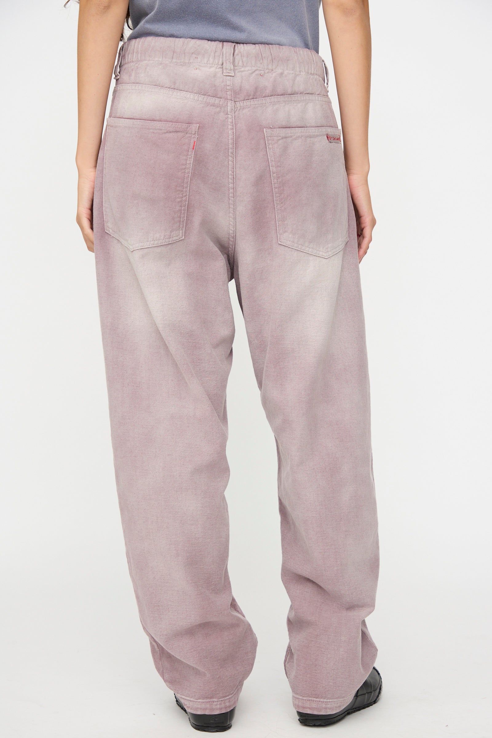 A person is seen from the back wearing Dr. Collectors’ P40 Z Boys Pant in Sunfaded Dusty Orchid—a relaxed fit style featuring a loose straight leg cut, large back pockets, and an elastic waistband, paired with black shoes. The comfortable, faded dusty orchid jeans are perfect for everyday wear.