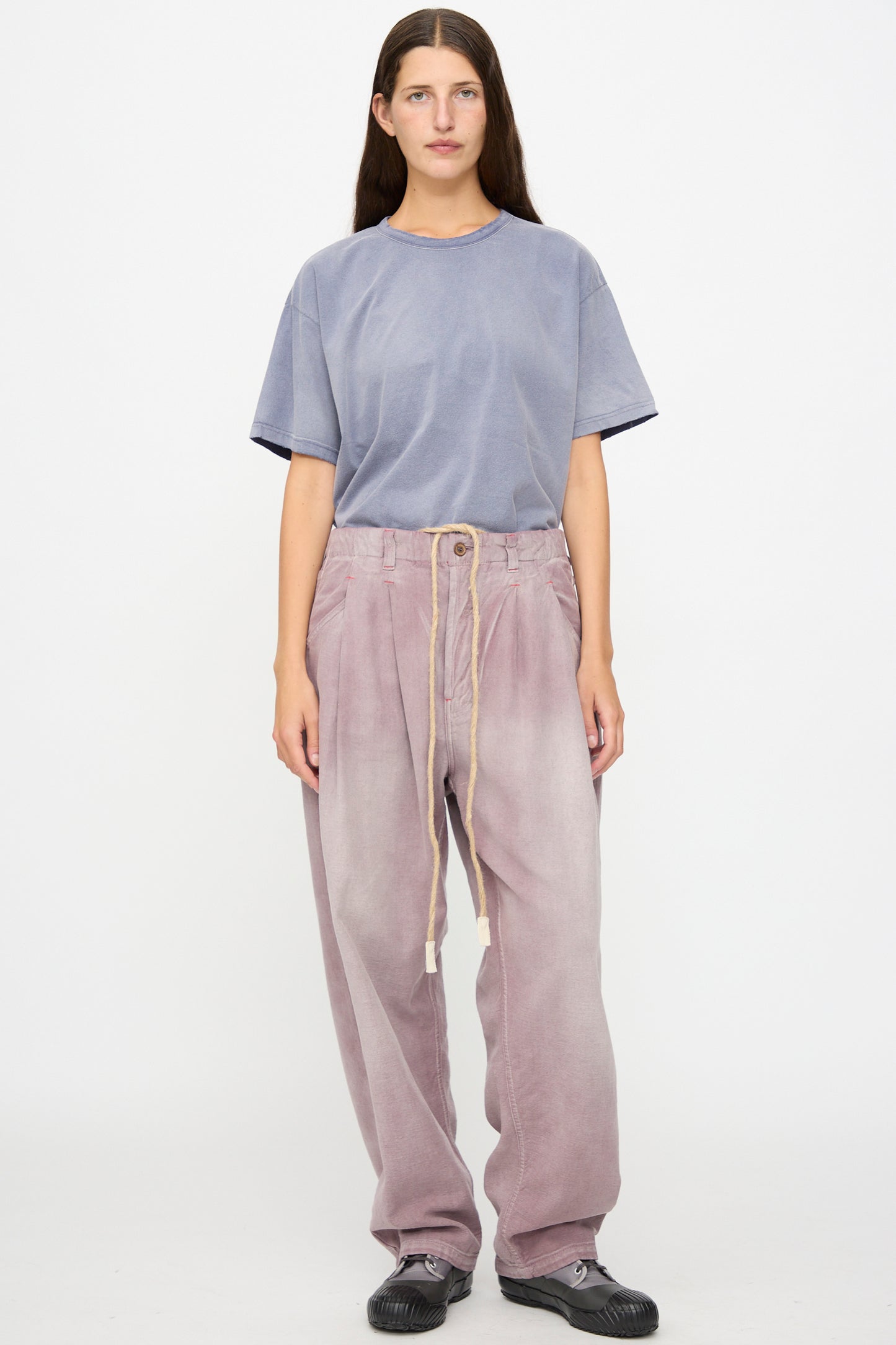 A person stands against a white background, wearing a loose-fitting blue t-shirt, Dr. Collectors P40 Z Boys Pant in Sunfaded Dusty Orchid—a relaxed fit pant crafted from light purple Japanese cotton with a drawstring—and black shoes.