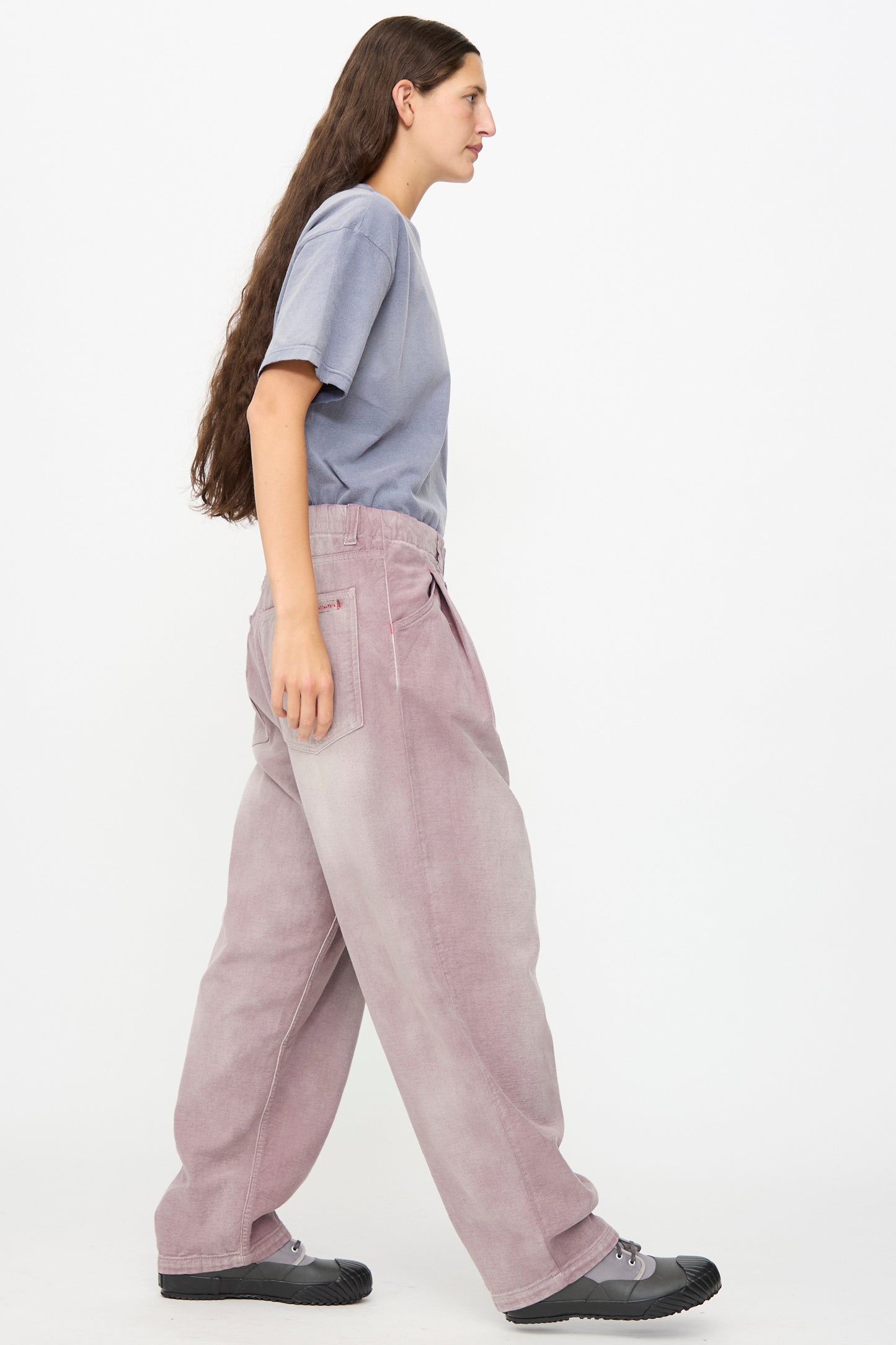 A person in a loose gray t-shirt, Dr. Collectors P40 Z Boys Pant in Sunfaded Dusty Orchid, and black shoes walks to the left against a plain white background.