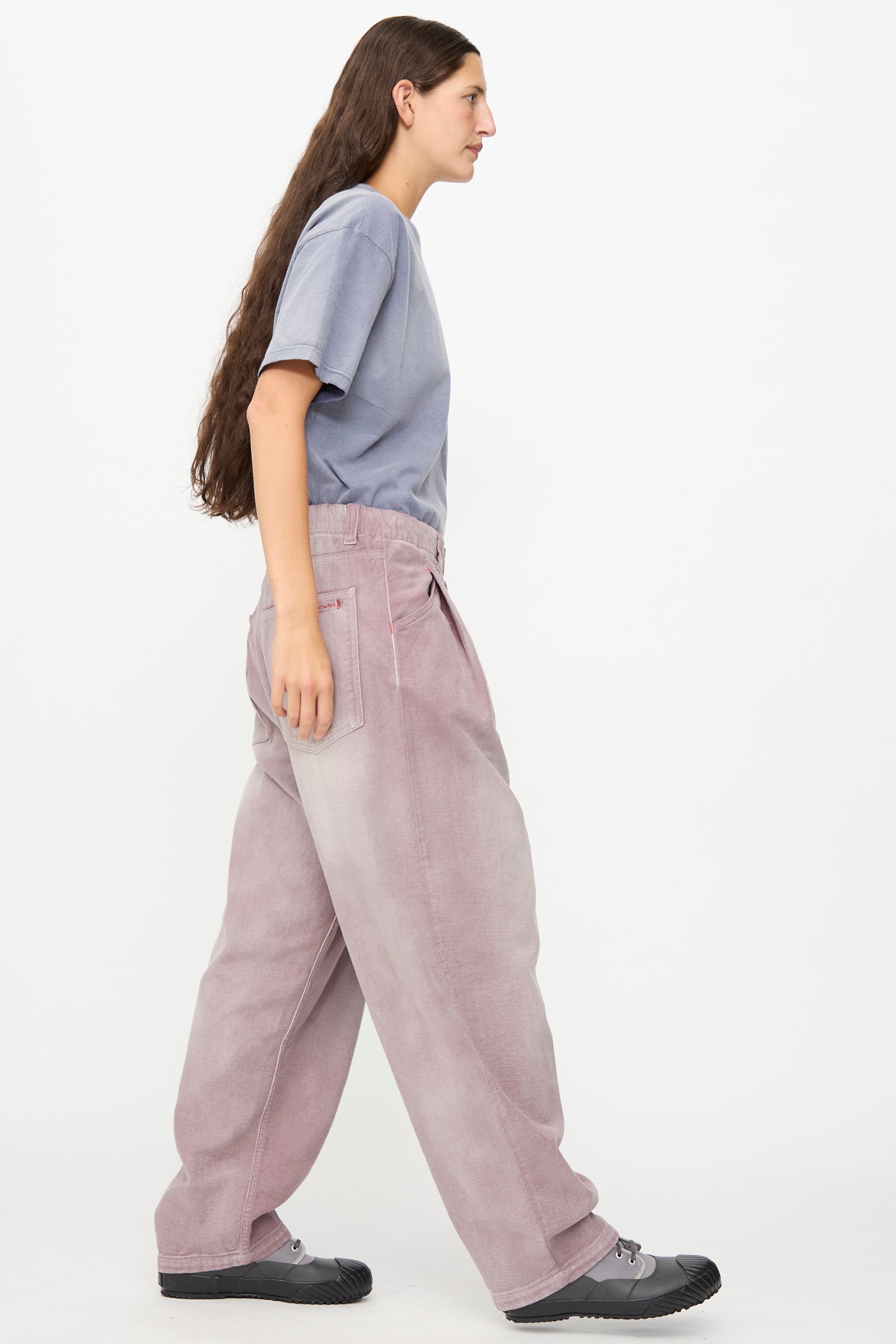 A person in a loose gray t-shirt, Dr. Collectors P40 Z Boys Pant in Sunfaded Dusty Orchid, and black shoes walks to the left against a plain white background.