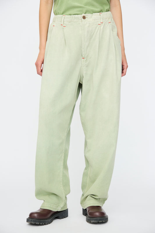 A person wears Dr. Collectors' P40 Z Boys Sunfaded Dye pants in Basil, paired with brown boots and a green shirt, standing against a plain white background.