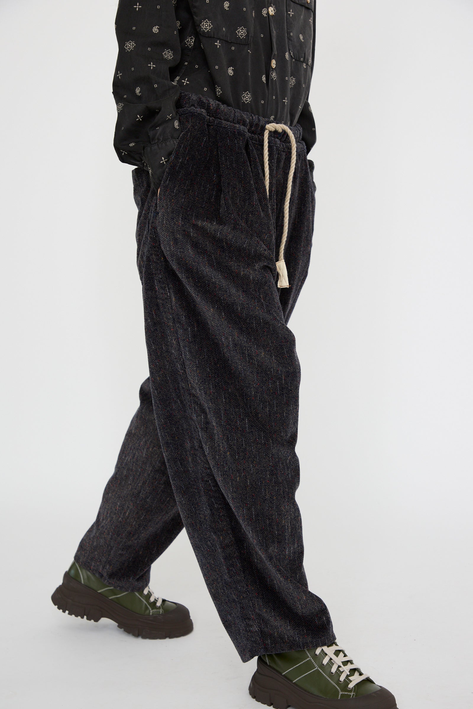 Wearing Dr. Collectors' P43 Kyoto Corduroy Japanese Dobby Tweed Pant in Black, a person pairs them with a patterned shirt and strides confidently wearing green and black shoes against a white background. 