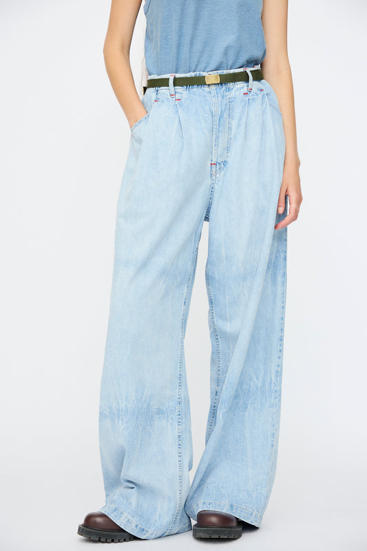 A person wearing a blue sleeveless top and Dr. Collectors P80 Venice Japanese Cotton Pant in Sky Blue stands against a plain white background, styled with an olive green belt and black shoes.