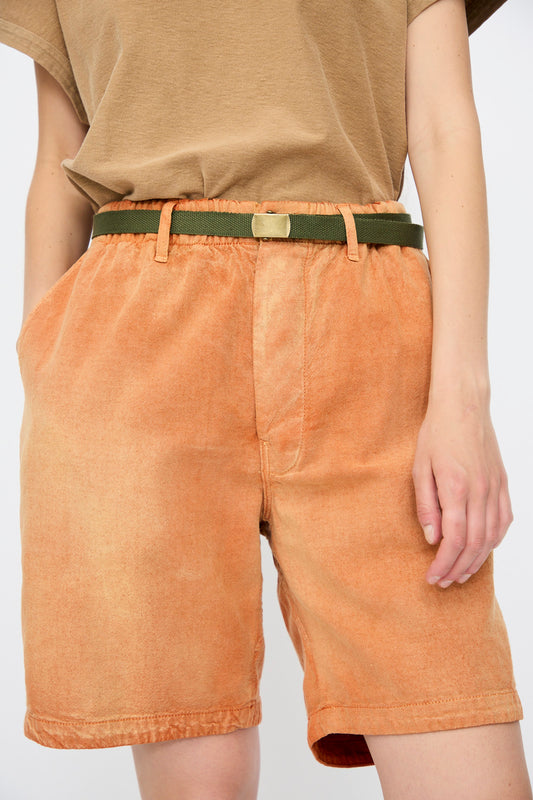 A person wears a tan t-shirt with Dr. Collectors P81 Honolulu Short Sunfaded in Rust, styled with a vintage military belt, hand in pocket, photographed from mid-torso to knees.