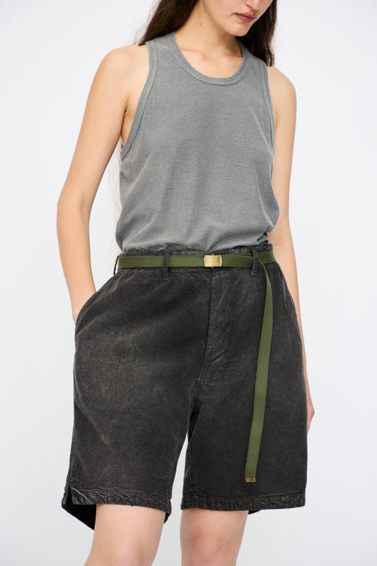 A person stands with hands in pockets against a plain background, wearing a gray sleeveless top and Dr. Collectors P81 Honolulu Short Sunfaded in Smoke Black, finished with a vintage military belt.