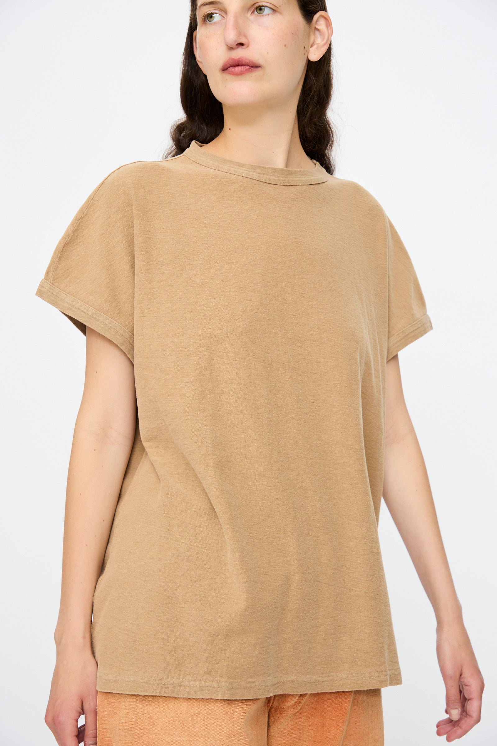Wearing the Dr. Collectors Palm Springs Tee Sunfaded in Mud—an organic cotton, loose-fit shirt made with plant-based dyes—a person stands against a plain white background, paired with light orange shorts.