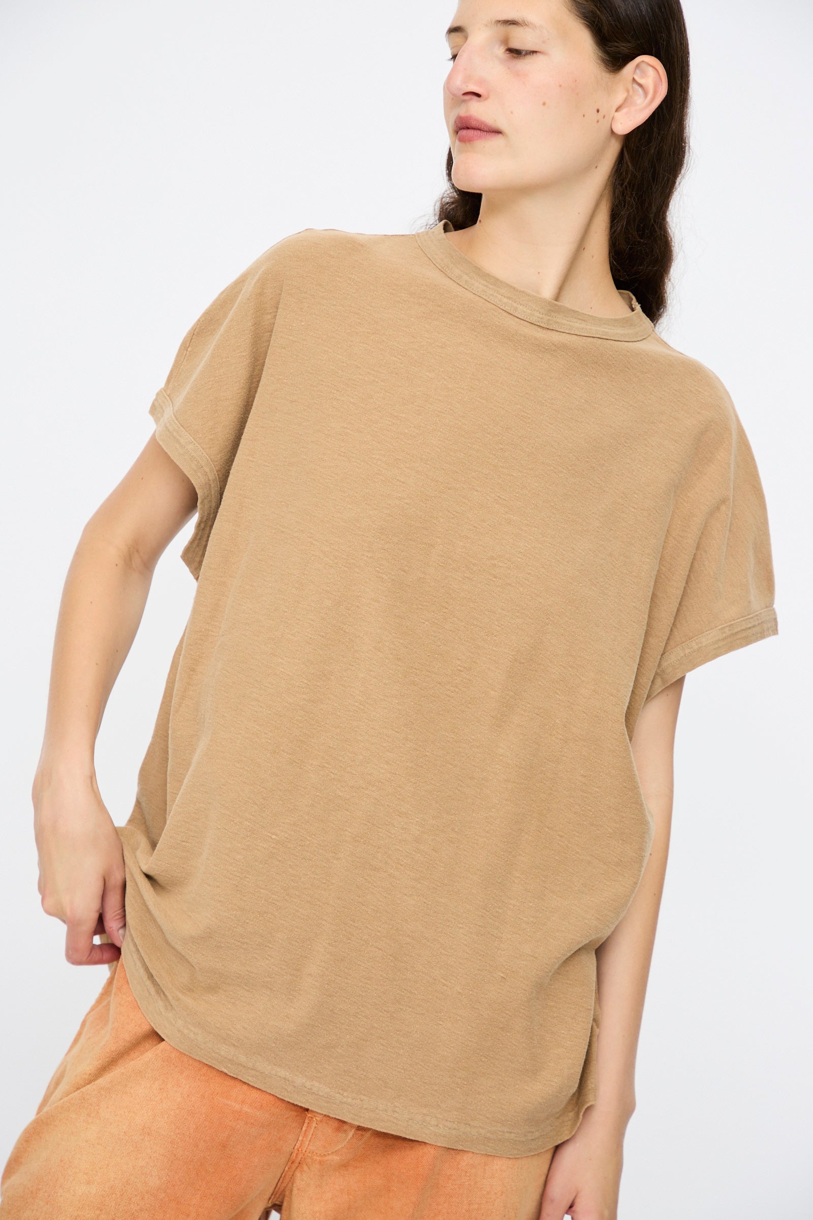 A person wears the Dr. Collectors Palm Springs Tee Sunfaded in Mud—an organic cotton, loose-fitting, short-sleeve top—paired with light orange pants against a plain light background.