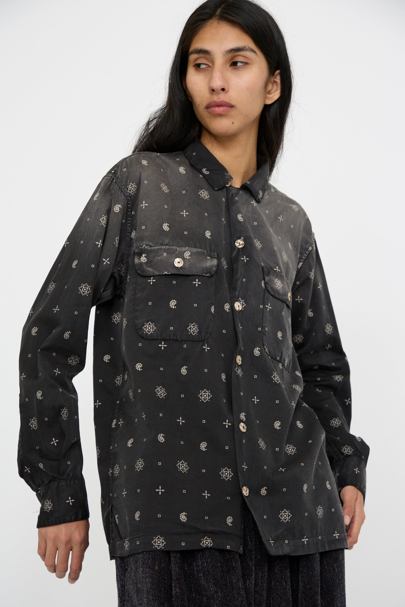 A person dressed in a Smoke Black Sateen Cotton Bowling Long Sleeve shirt by Dr. Collectors, featuring patterned fabric and abalone shell buttons, gazes to the side.