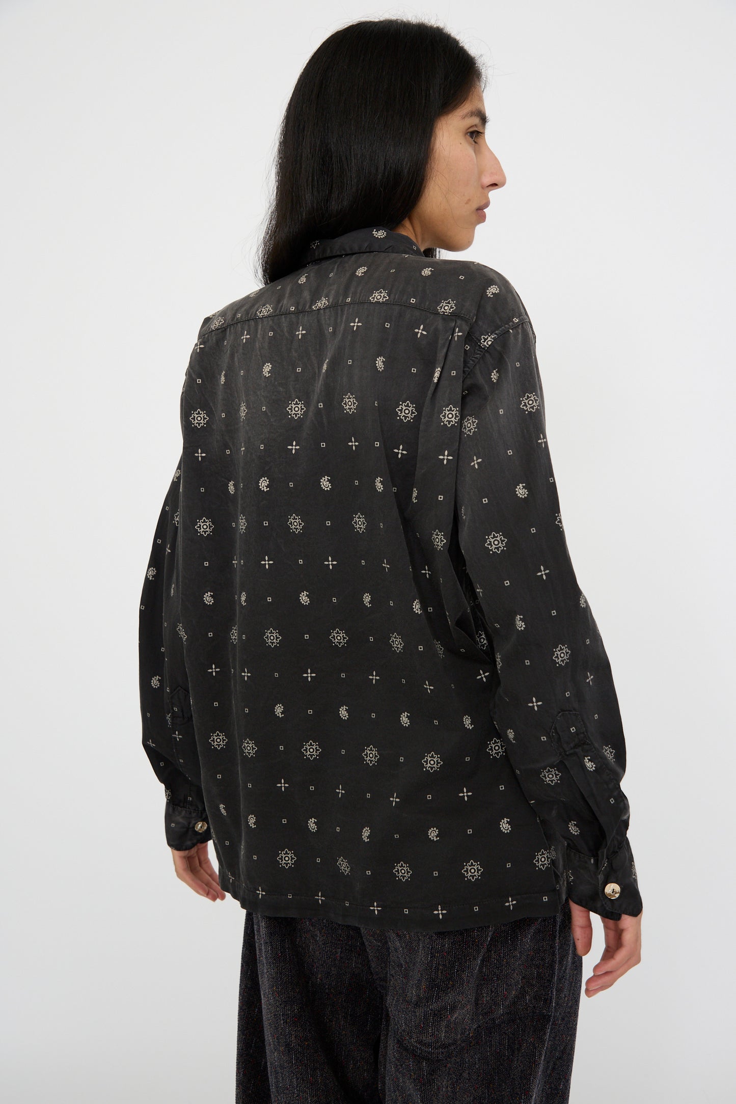 A person with flowing long hair models the stylish Sateen Cotton Bowling Long Sleeve in Smoke Black by Dr. Collectors, adorned with Abalone shell buttons, as they stand sideways against a plain background.