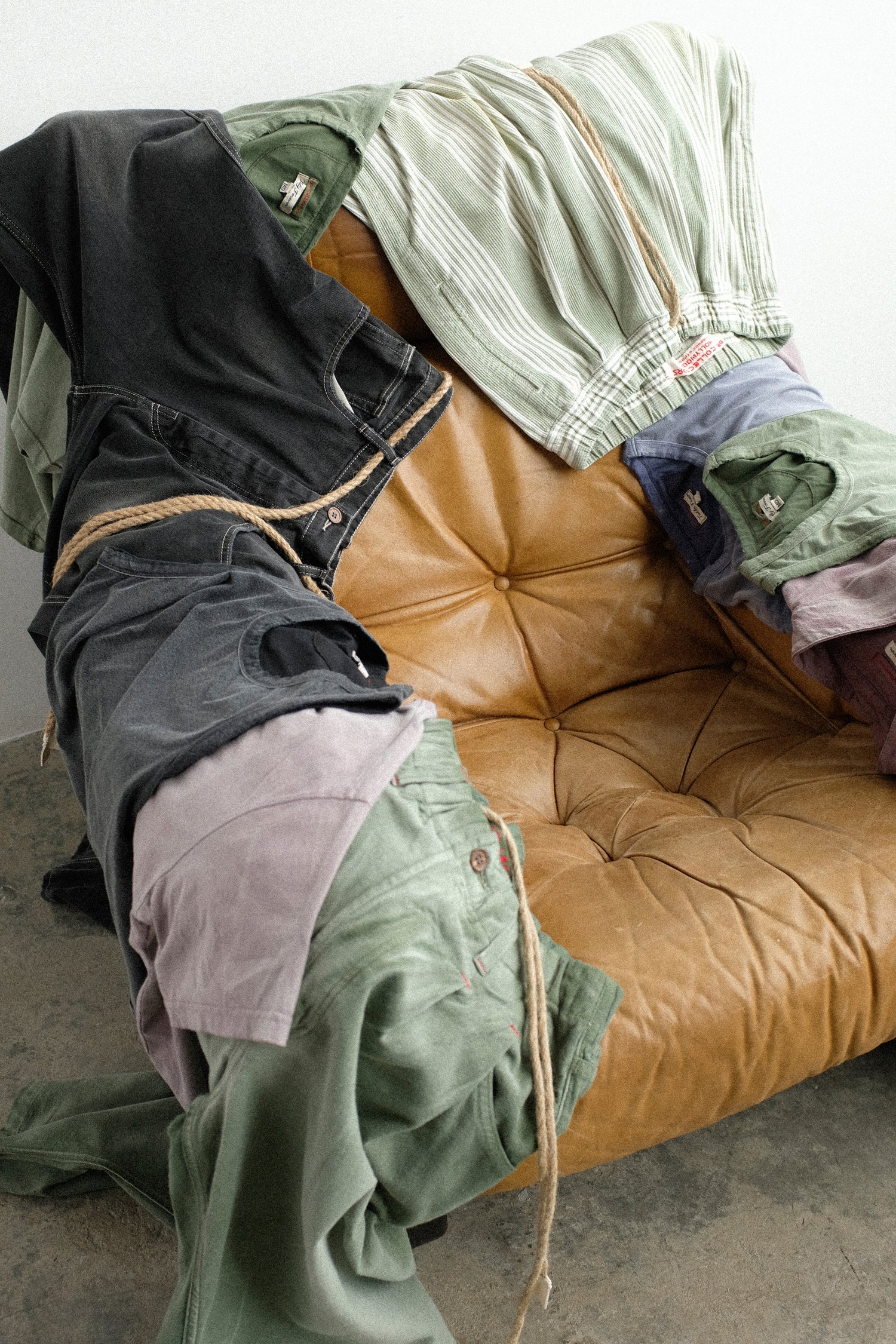 A pile Dr. Collectors clothing, including green striped pants, a black denim pants, and olive green shirts, draped over a tufted tan leather armchair in a studio setting.