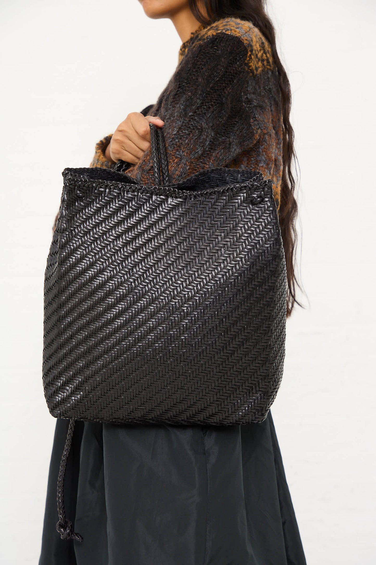 A person holds a Dragon Diffusion Backpack in Black, featuring traditional braided weaving and a shoulder strap. The person, dressed in a dark, textured top and sporting long brown hair, showcases the handcrafted bag with the focus on the intricate design of the bag and their torso.