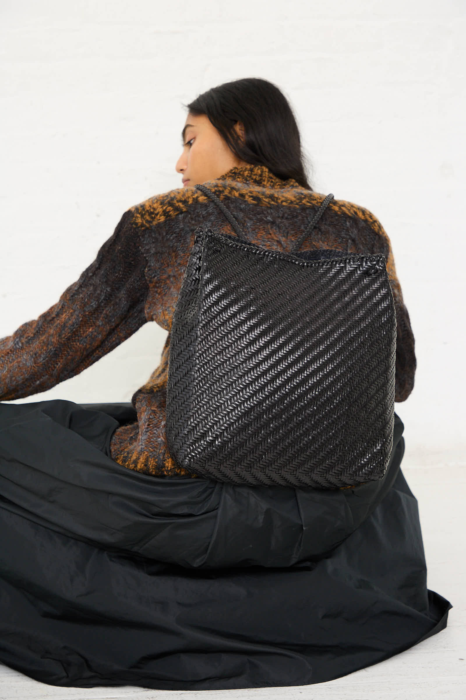 A person is sitting with their back to the camera, wearing a textured, dark-colored sweater, a black skirt, and carrying the Dragon Diffusion Backpack in Black.