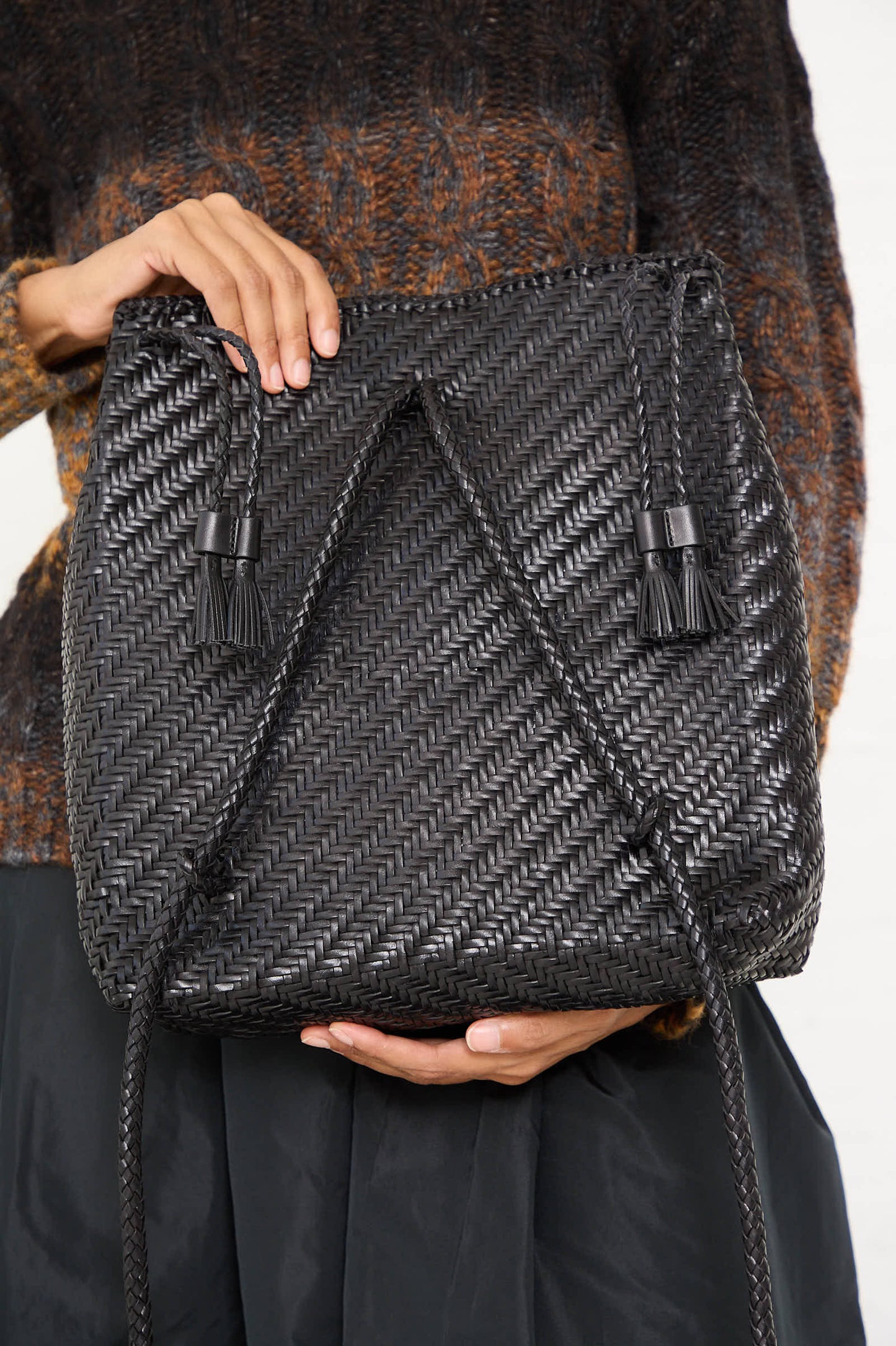 Person holding a Dragon Diffusion Backpack in Black with drawstrings and tassels against their torso, wearing a textured sweater and dark skirt. The bag's detailed design showcases traditional braided weaving, adding an artisanal touch to the look.