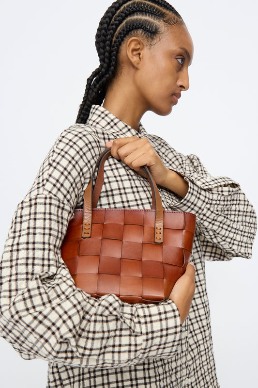 A person with braided hair wears a plaid shirt and holds the Dragon Diffusion Japan Mini Tote in Tan, a small basket-shaped cow leather bag, against a plain background.