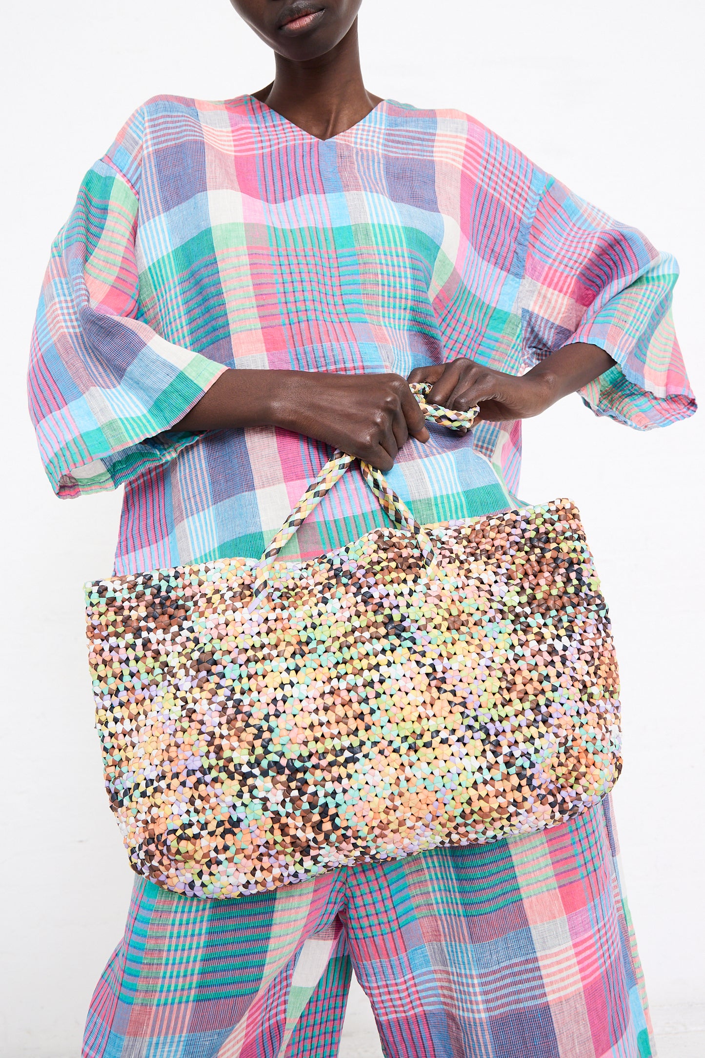A woman holding a large Octo Multi in Pastel hand-knotted leather tote from Dragon Diffusion, wearing a colorful plaid blouse with voluminous sleeves, against a white background.