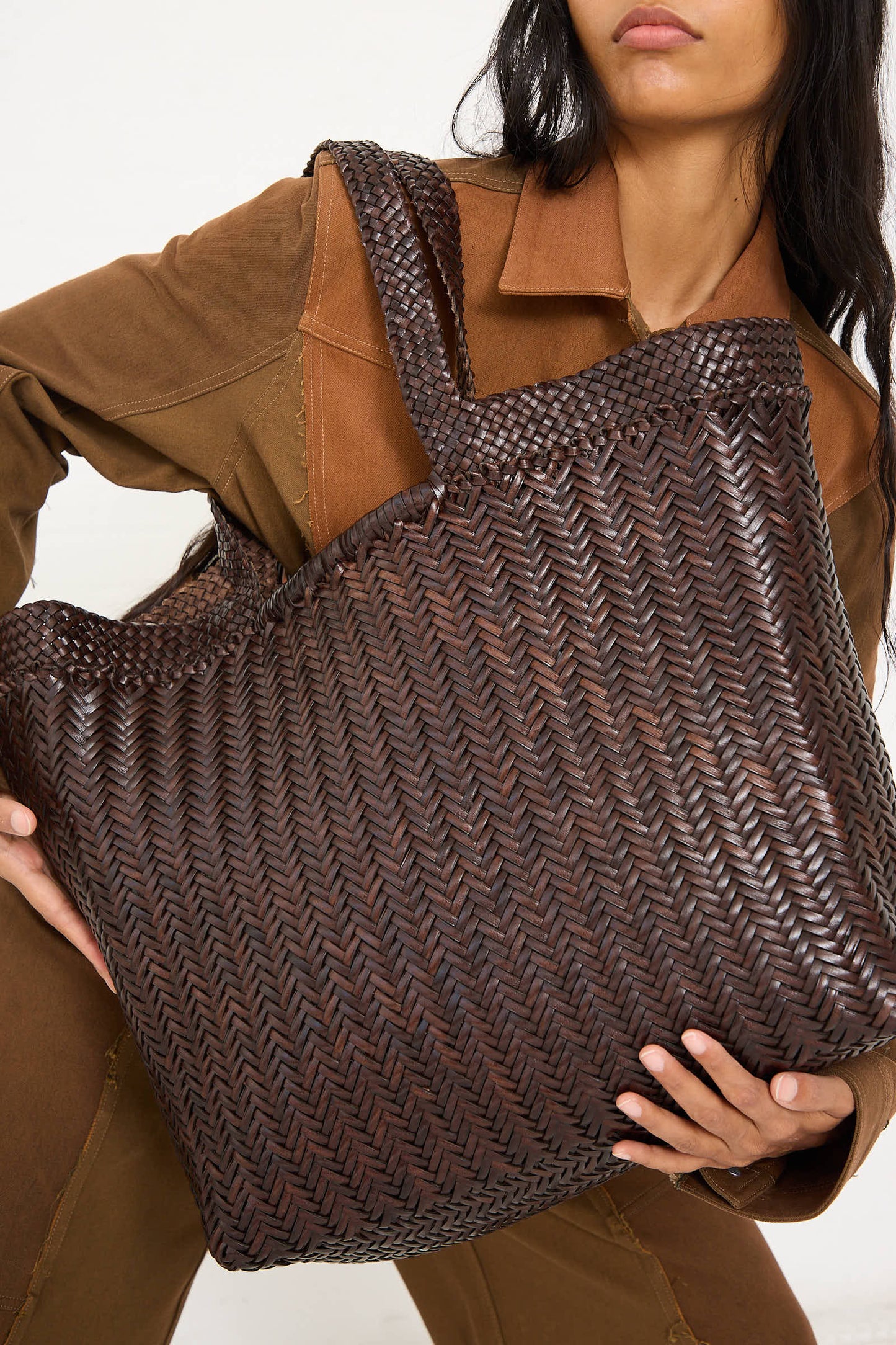 A person in brown clothing holds the Dragon Diffusion Oversize Triple Jump Weave Maxi Shopper in Dark Brown, skillfully crafted using traditional weaving techniques.