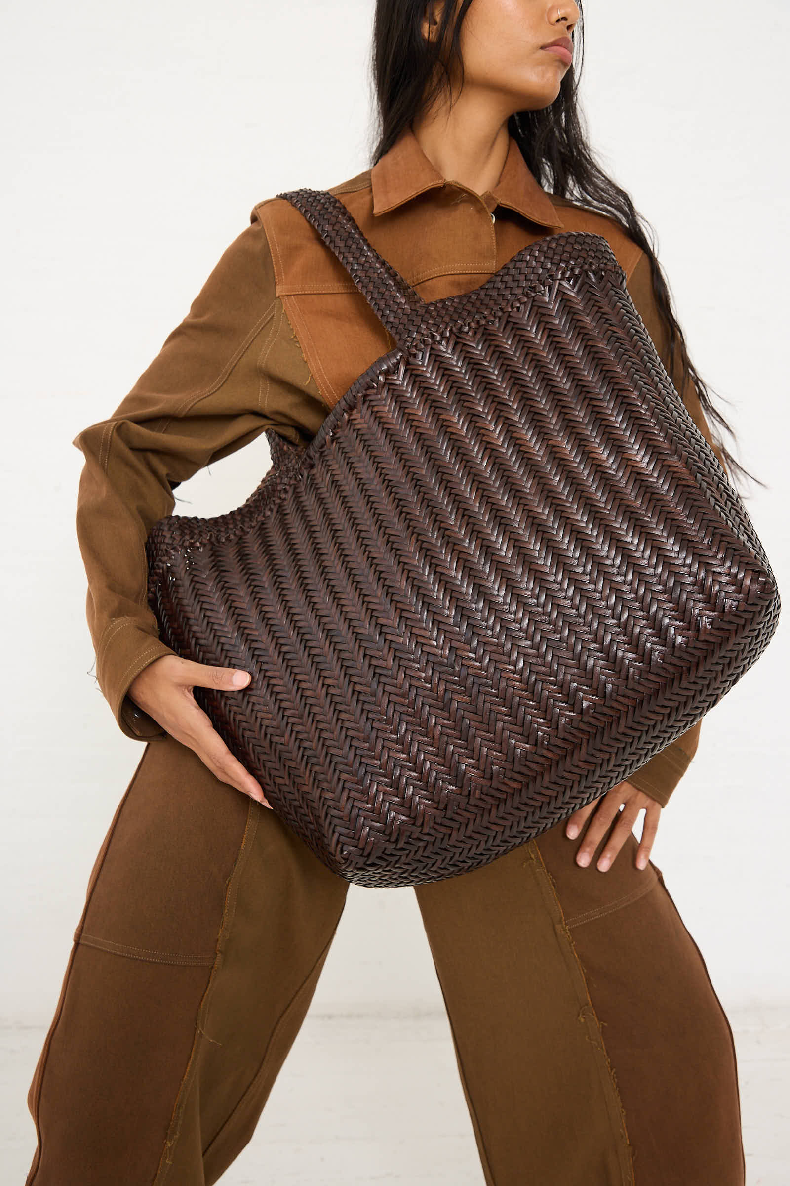 Person holding the Dragon Diffusion Oversize Triple Jump Weave Maxi Shopper in Dark Brown, set against a neutral backdrop and wearing a brown two-piece outfit.