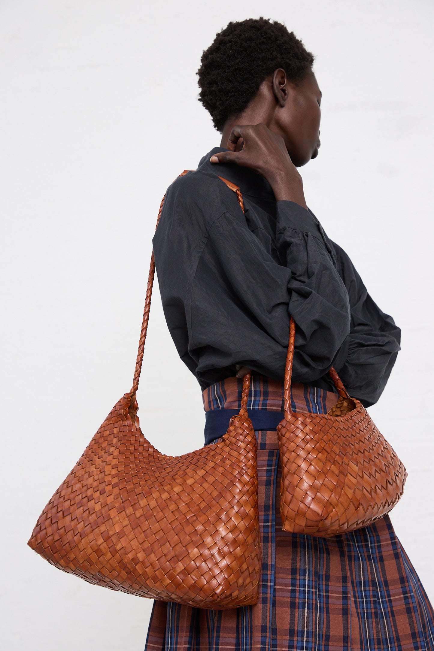 A woman in profile wearing a dark blouse and plaid skirt, holding two large handwoven Dragon Diffusion Rosanna in Tan bags, against a white background.