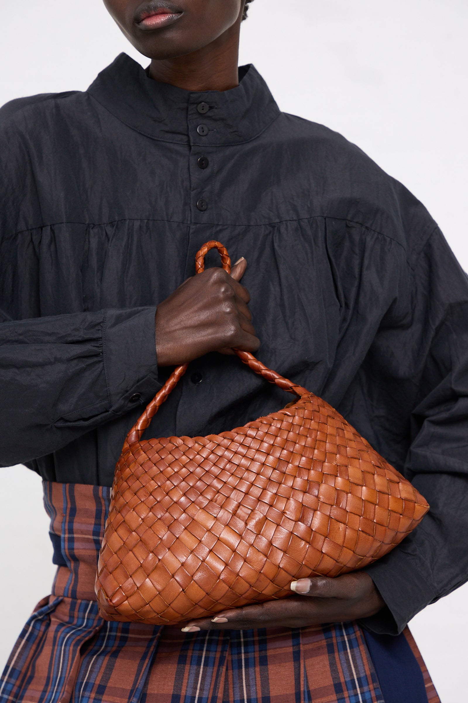 A person in a dark ruffled shirt and plaid pants holding a Dragon Diffusion handwoven brown leather bag called Rosanna in Tan. Only the torso and hands are visible.