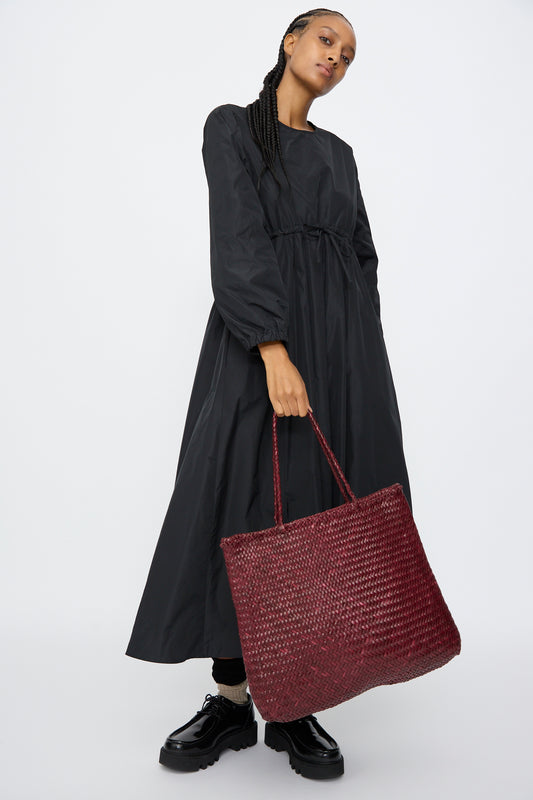 A person holds the Dragon Diffusion Sophie Large bag in Bordo, styled with a long black dress and black shoes, against a plain white background.