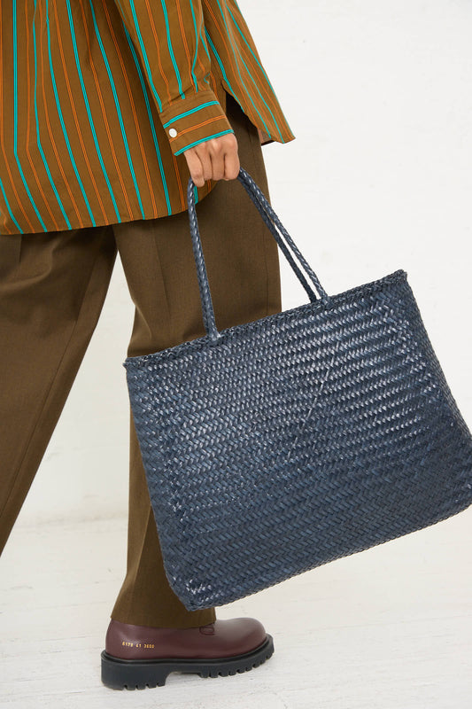 Person carrying a Dragon Diffusion Sophie Large Tote in Marine, showcasing intricate braided weaving techniques, while wearing a brown and green striped shirt, brown pants, and brown shoes.