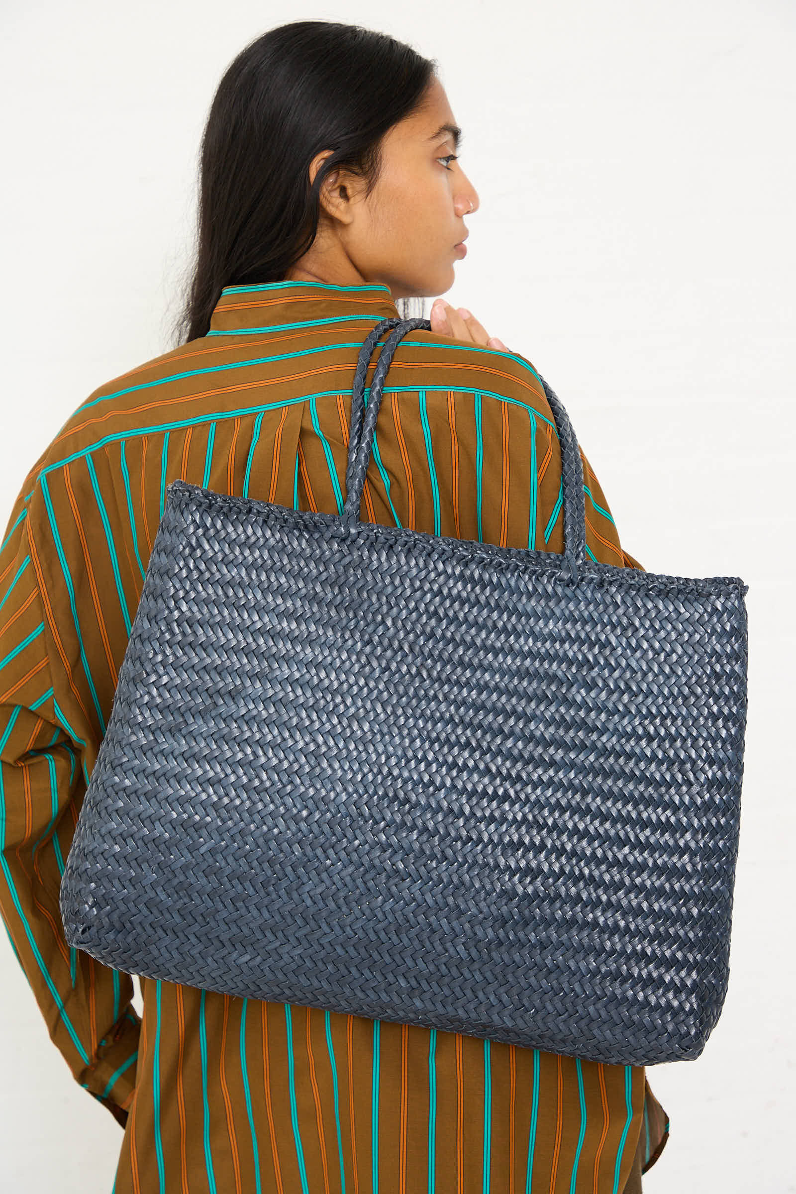 A person with long dark hair, wearing a striped brown shirt, is holding the Sophie Large Tote in Marine by Dragon Diffusion over their shoulder.