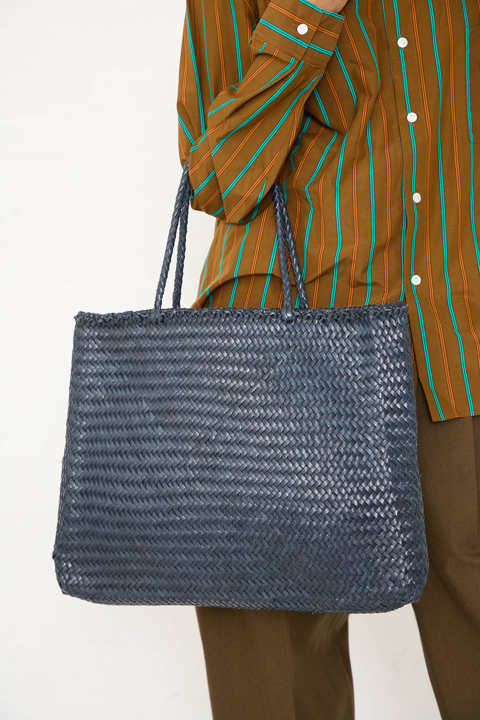 Person holding a Dragon Diffusion Sophie Large Tote in Marine with exquisite braided weaving techniques, wearing a striped brown shirt and brown pants.