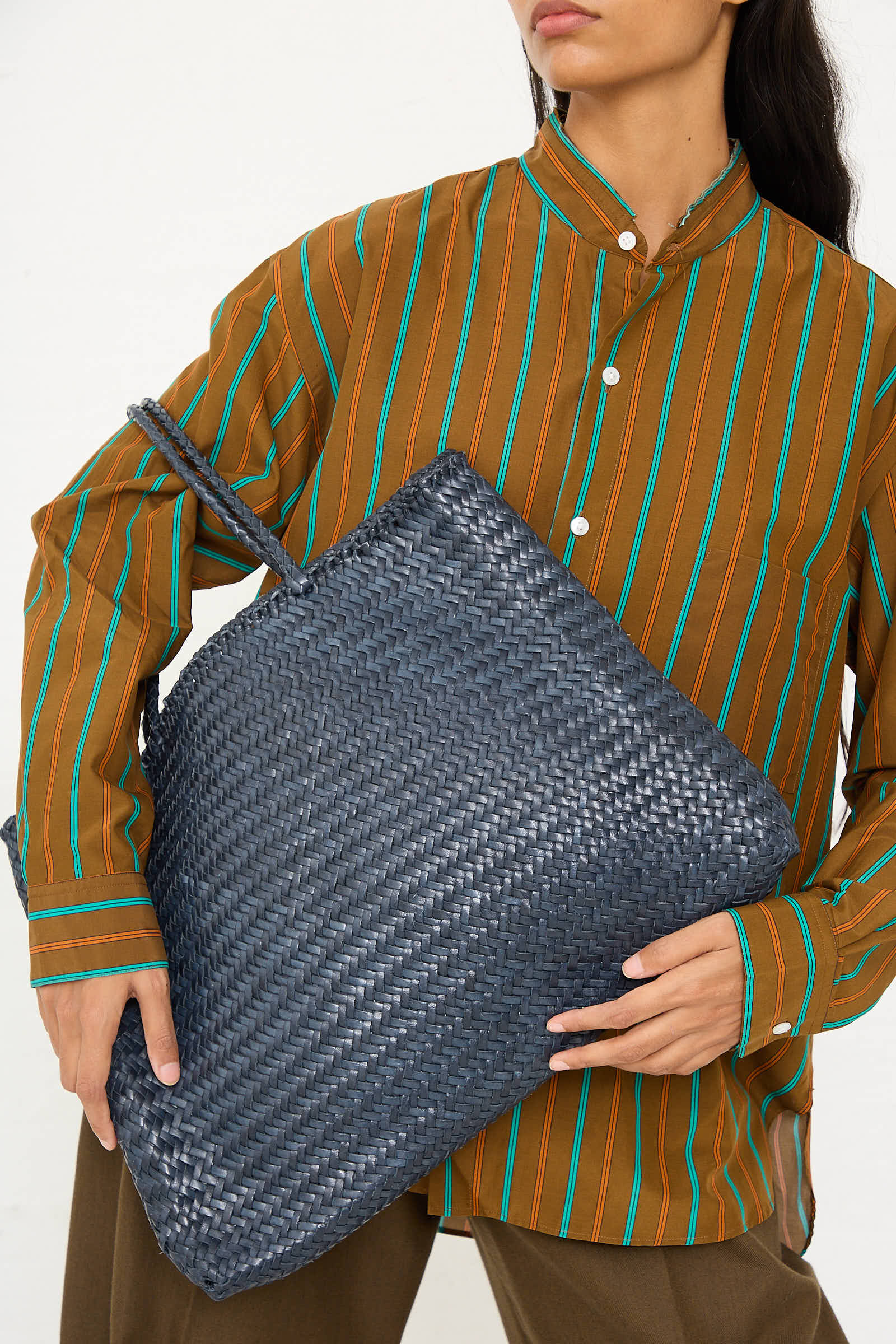 A person wearing a brown striped shirt holds the Sophie Large Tote in Marine by Dragon Diffusion against their side, showcasing its intricate braided weaving techniques.