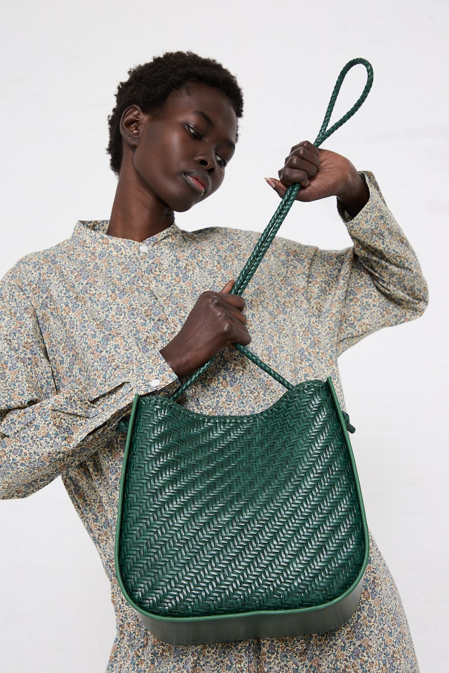 A woman examining a Dragon Diffusion Wanaka in Forest handwoven leather handbag, dressed in a floral print outfit, against a white background.