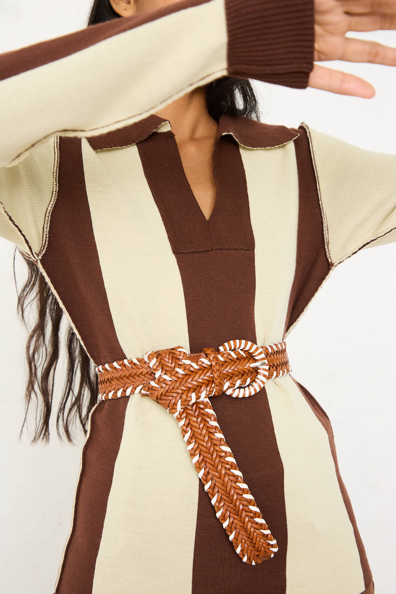 A person wearing a long-sleeve, brown and beige striped dress paired with the Wrapped Leather Buckle Belt in Tan and White by Dragon Diffusion. The person's arms are partially raised, obscuring part of their face.