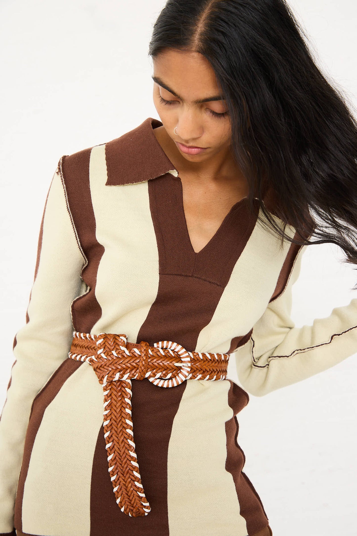 A woman with long, dark hair wears a beige and brown striped sweater dress featuring a V-neck, accessorized with the Wrapped Leather Buckle Belt in Tan and White by Dragon Diffusion. She is looking down and to her side.