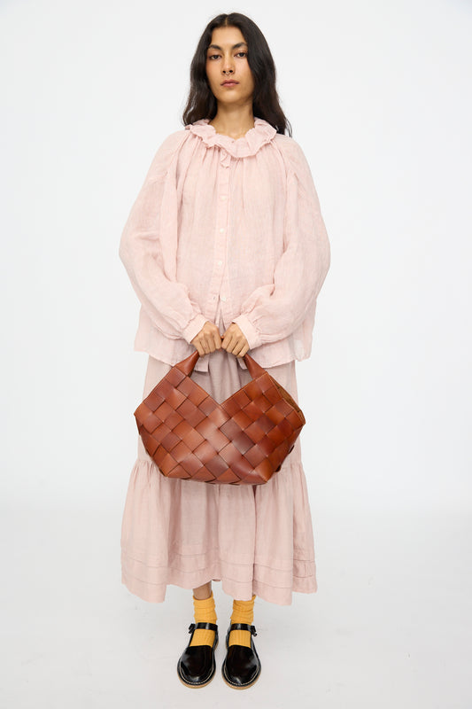 A woman stands against a plain background wearing a loose light pink dress, yellow socks, black shoes, and holding the Dragon Diffusion Botteghe Bag in Tan.