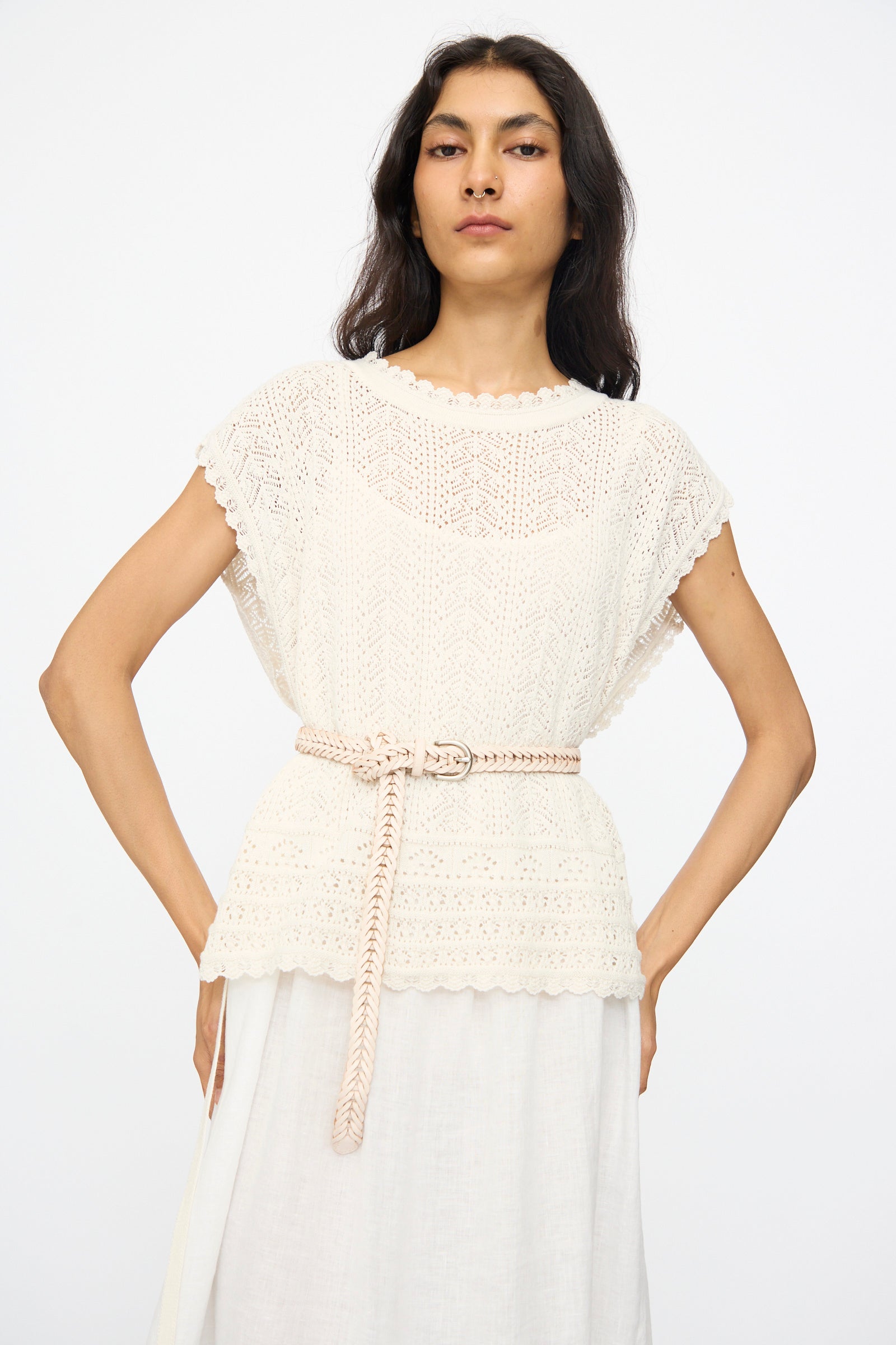 A woman stands against a plain white background wearing a cream crochet short-sleeve top with a scalloped hem, a white skirt, and the Brass Buckle Single Link Belt in Sand by Dragon Diffusion.